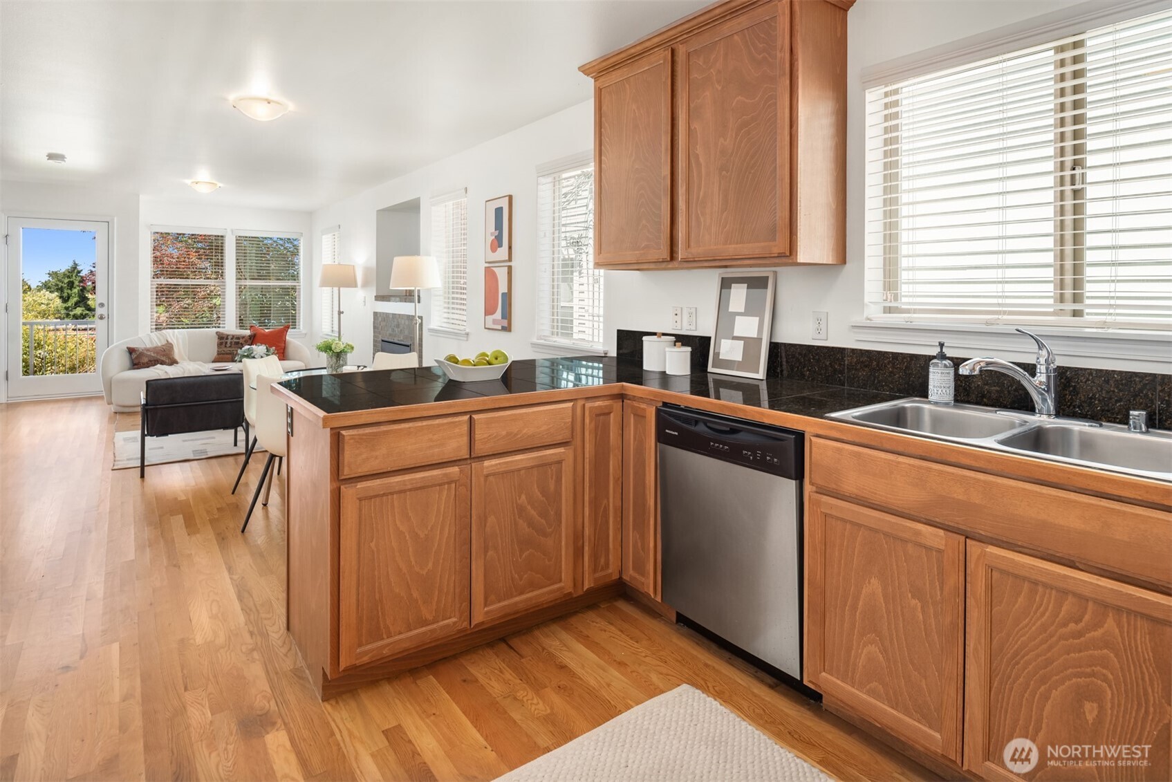 Open-plan kitchen with warm wood cabinets and black countertops, looking toward a bright living area and patio door.