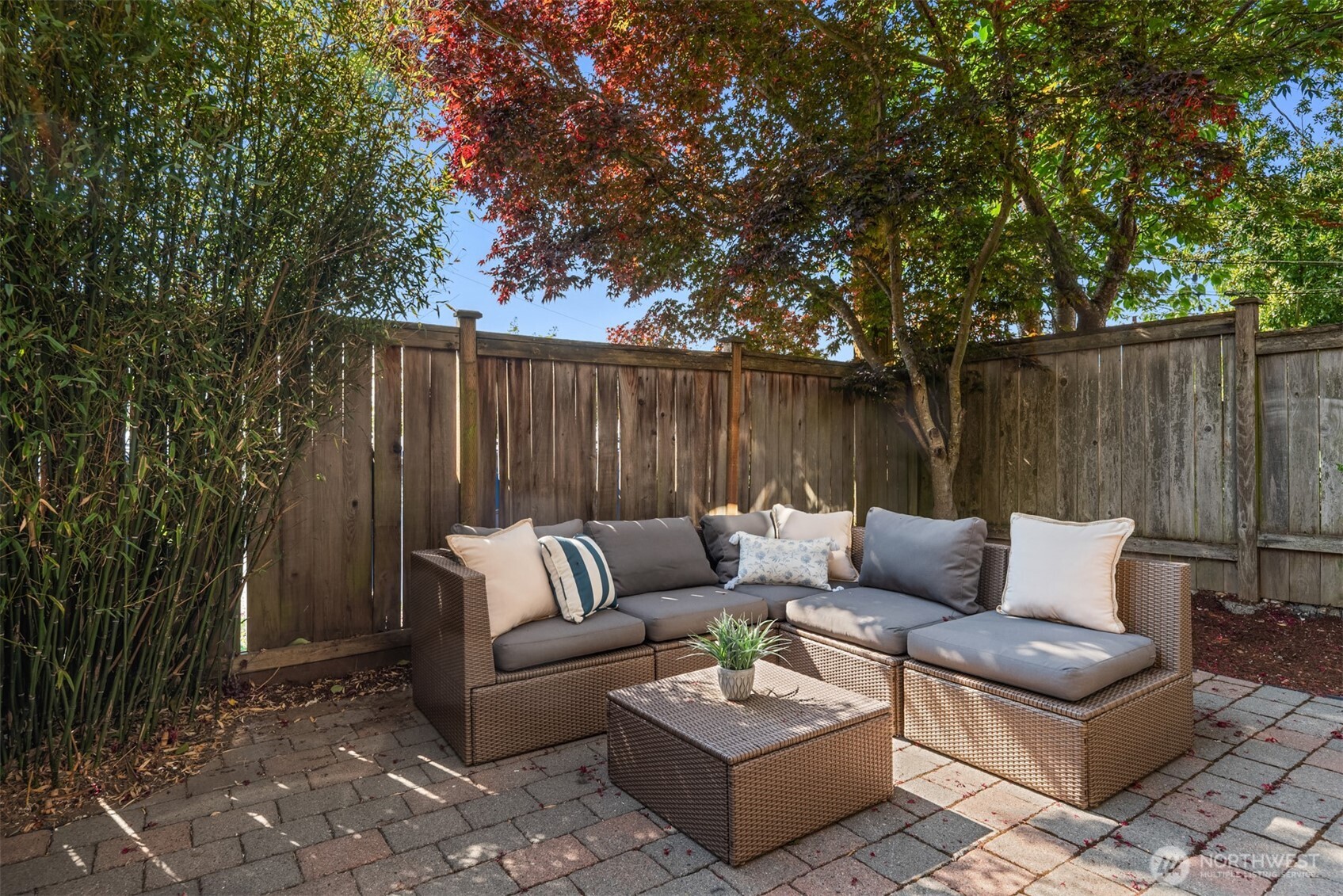 Wicker patio sectional and coffee table beneath trees, enclosed by a wooden privacy fence.