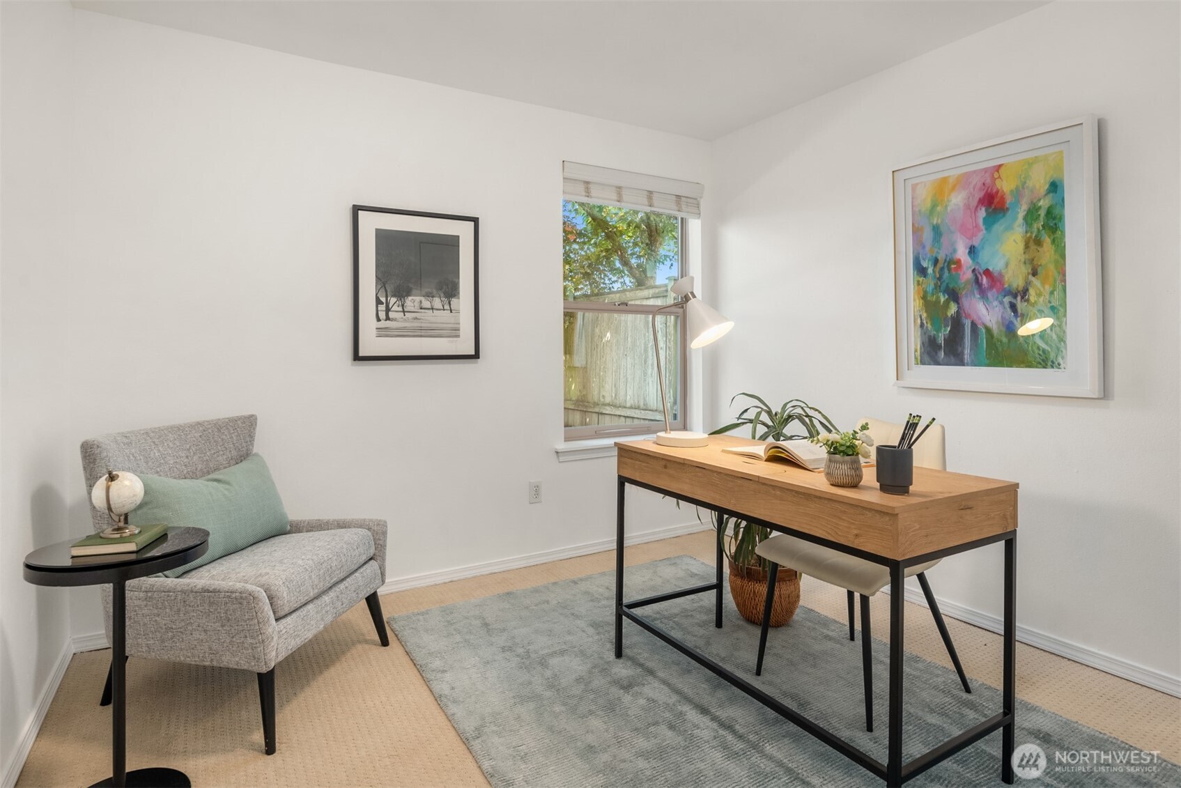 Furnished home office with a wooden desk, gray armchair, and colorful artwork.