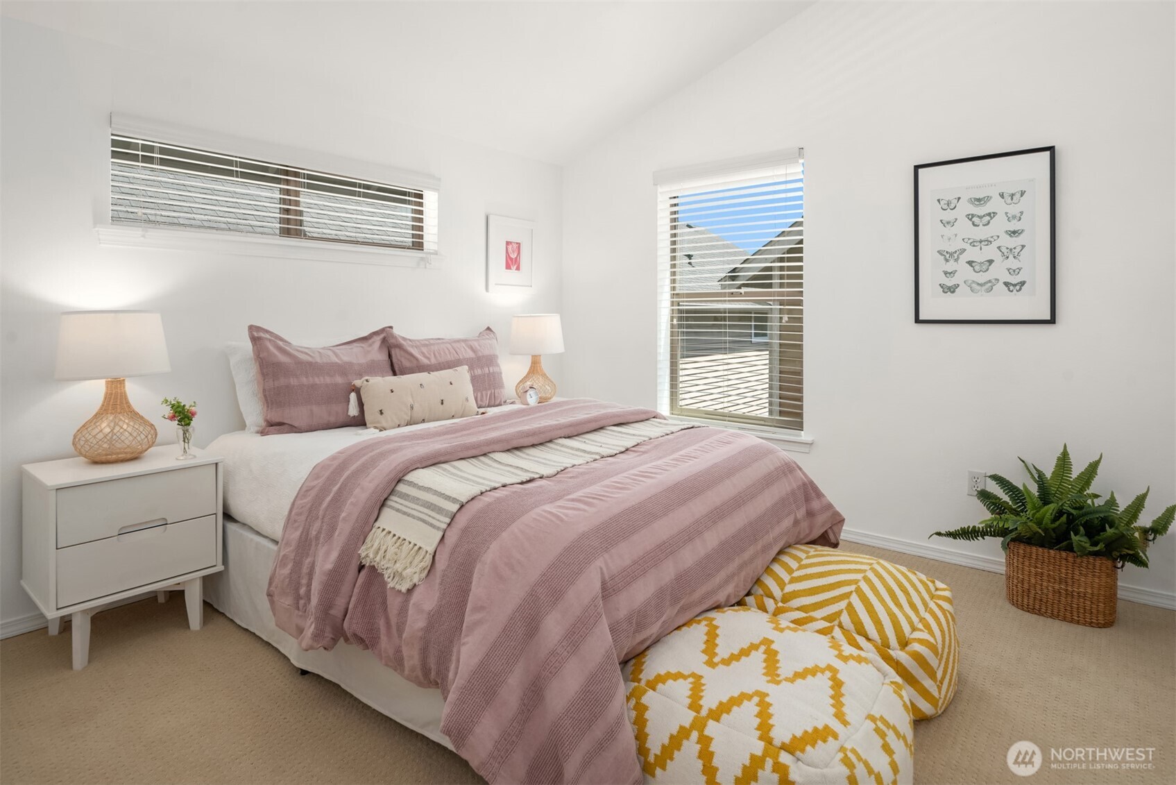 Staged bedroom with dusty pink bedding, mustard patterned floor cushions, and two windows with blinds.