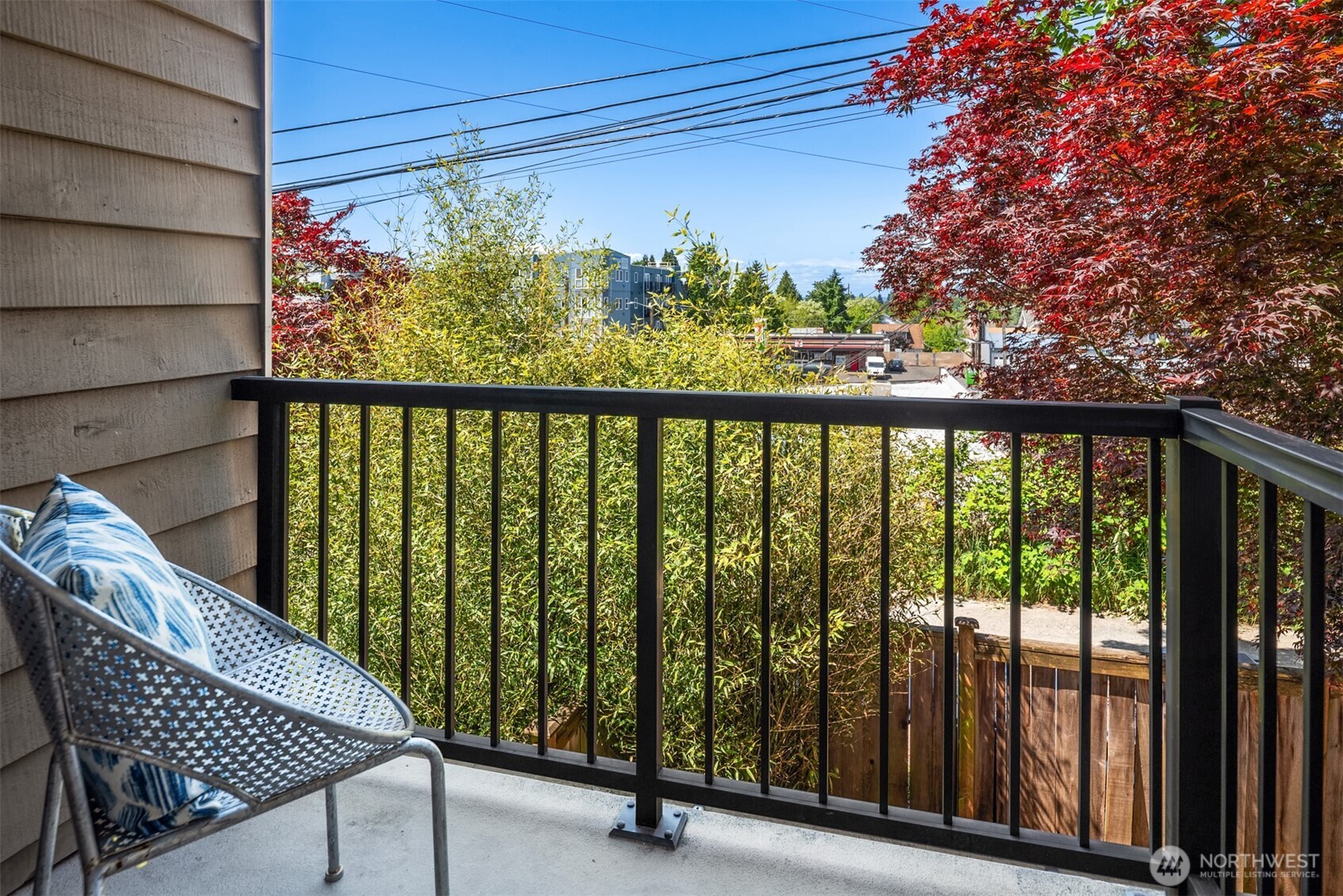 Balcony with a patterned chair, black railing, leafy trees, and nearby buildings.