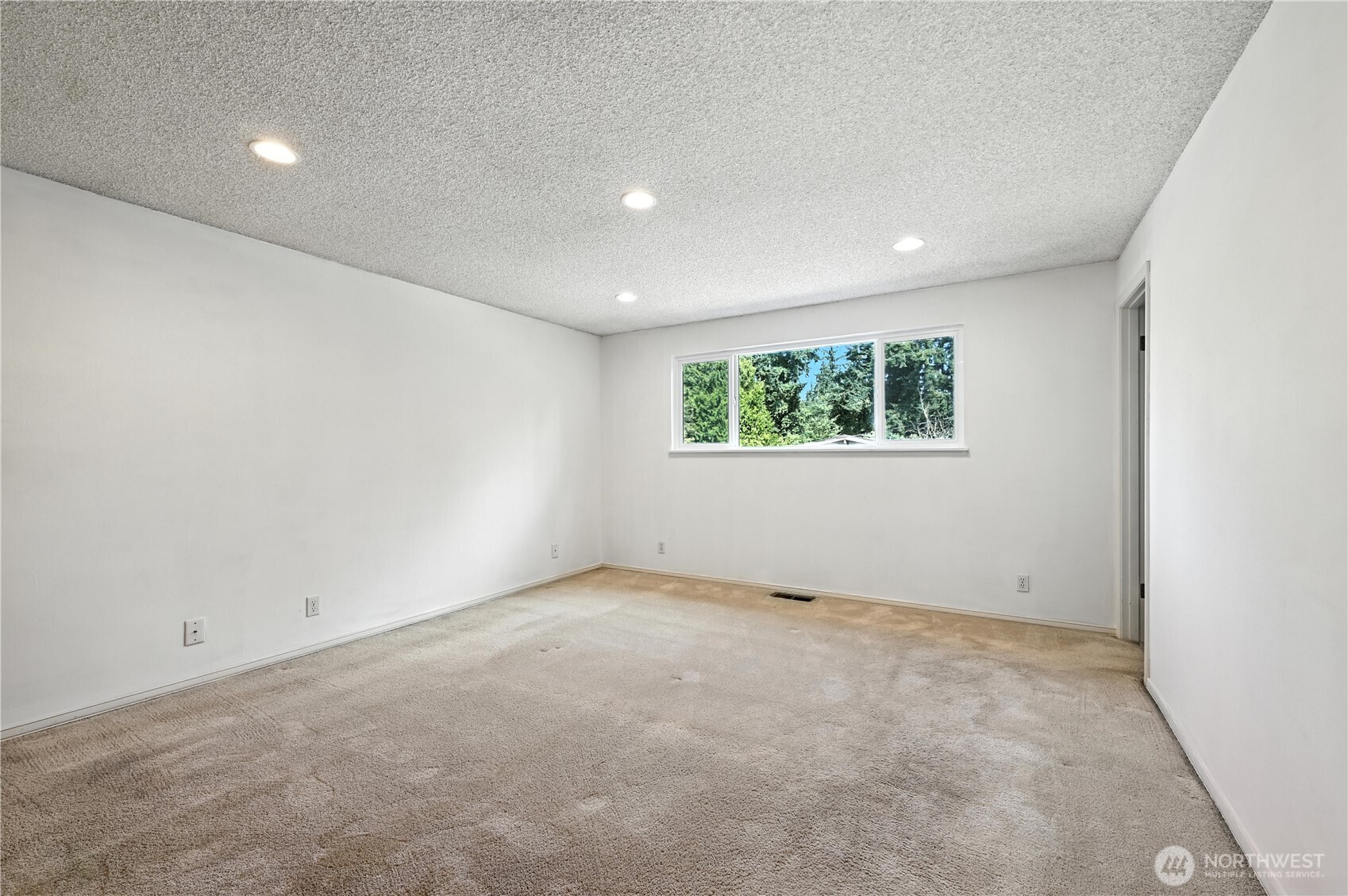 Empty carpeted room with white walls, recessed lights, and a wide window overlooking trees.