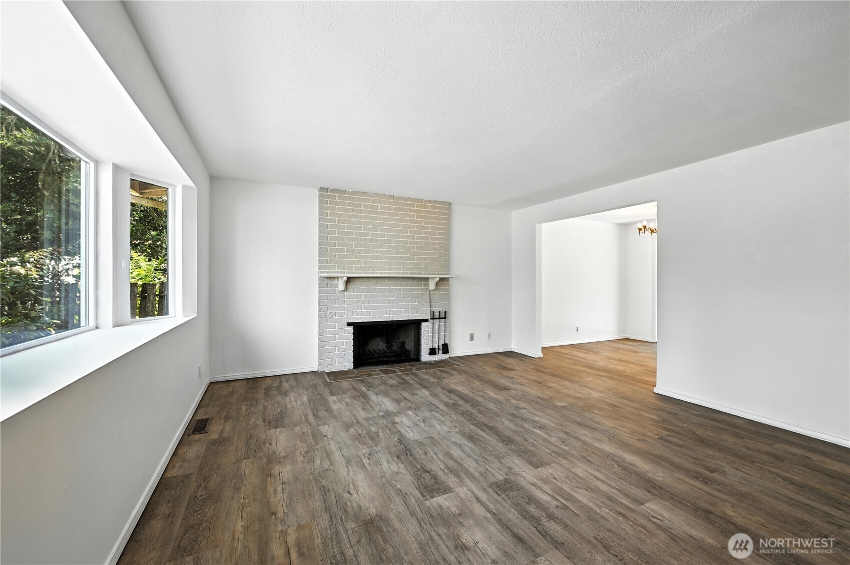 Vacant living room with a white brick fireplace, wide-plank flooring, and large windows.