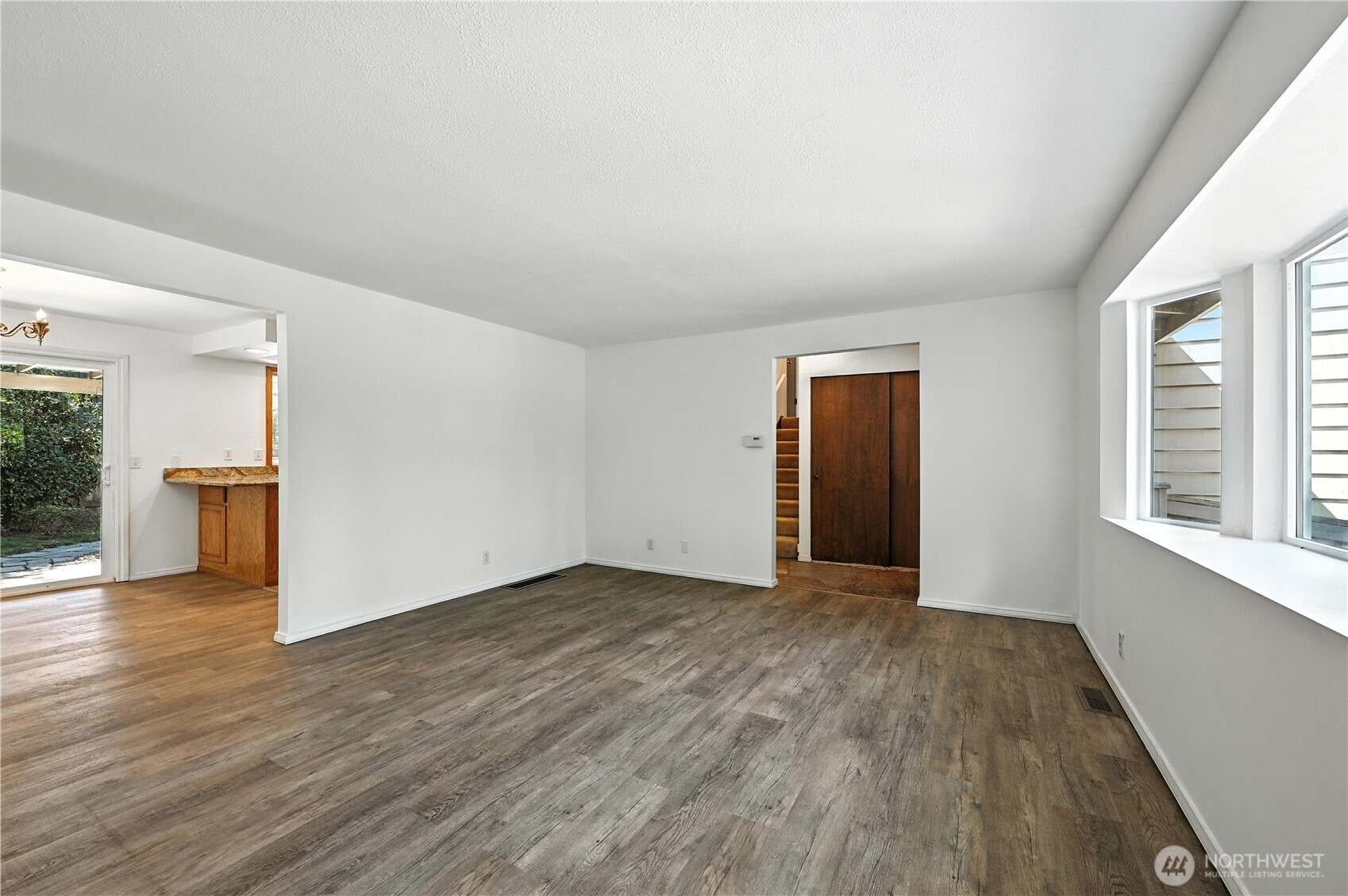 Empty living room with gray-brown plank flooring, broad windows, and a doorway to the entry.