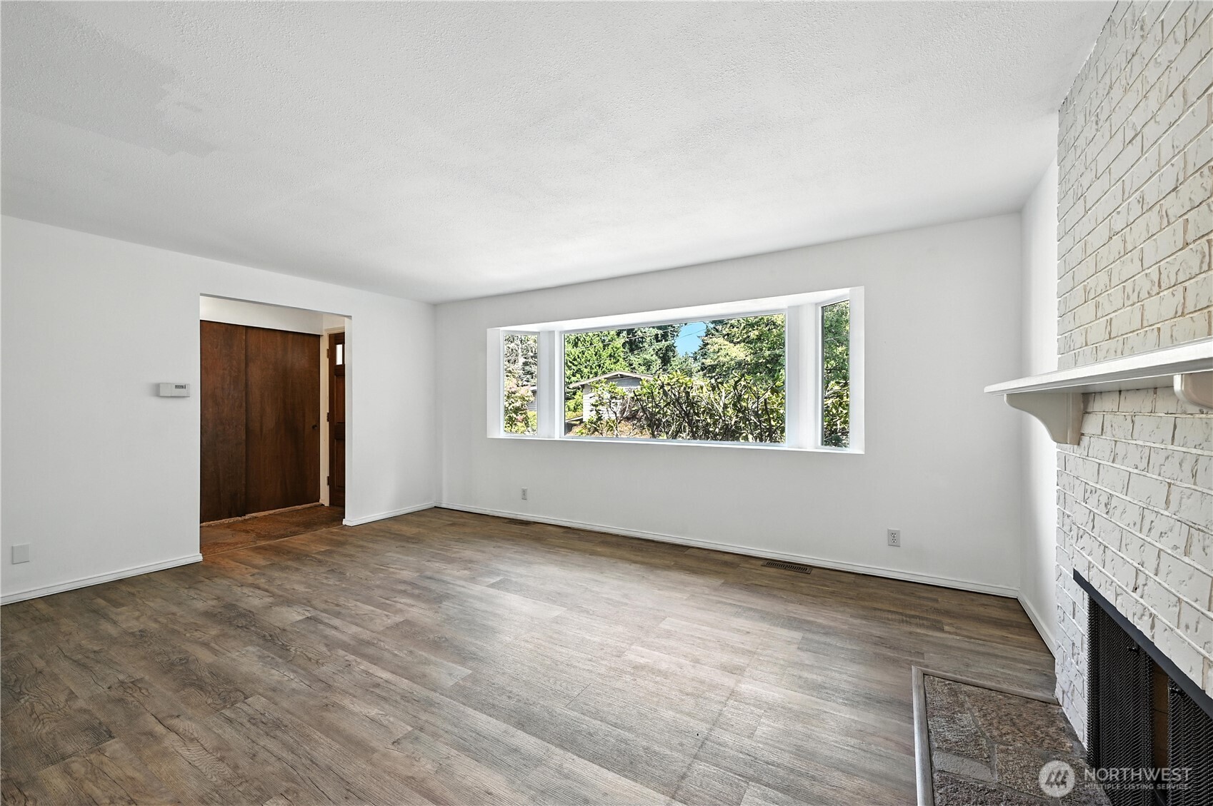 Empty living room with wood-look floors, a white brick fireplace, and a wide window overlooking greenery.