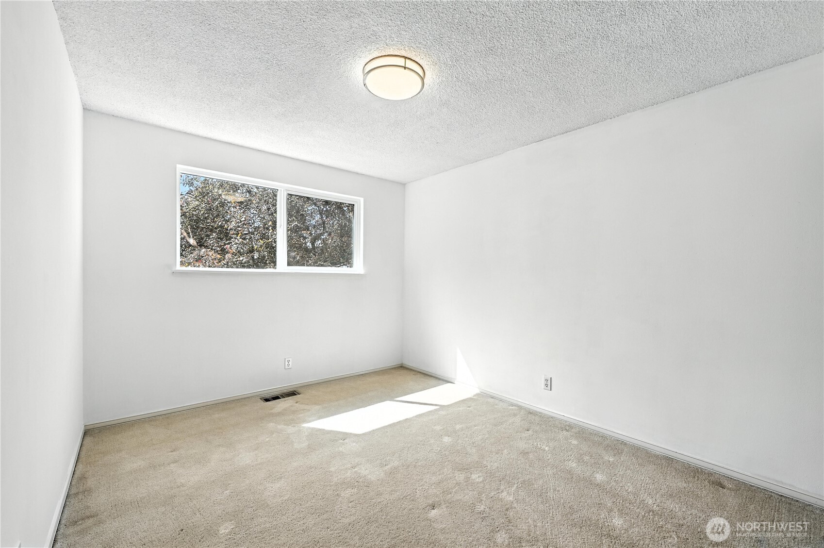 Empty carpeted room with white walls and a small window overlooking trees.