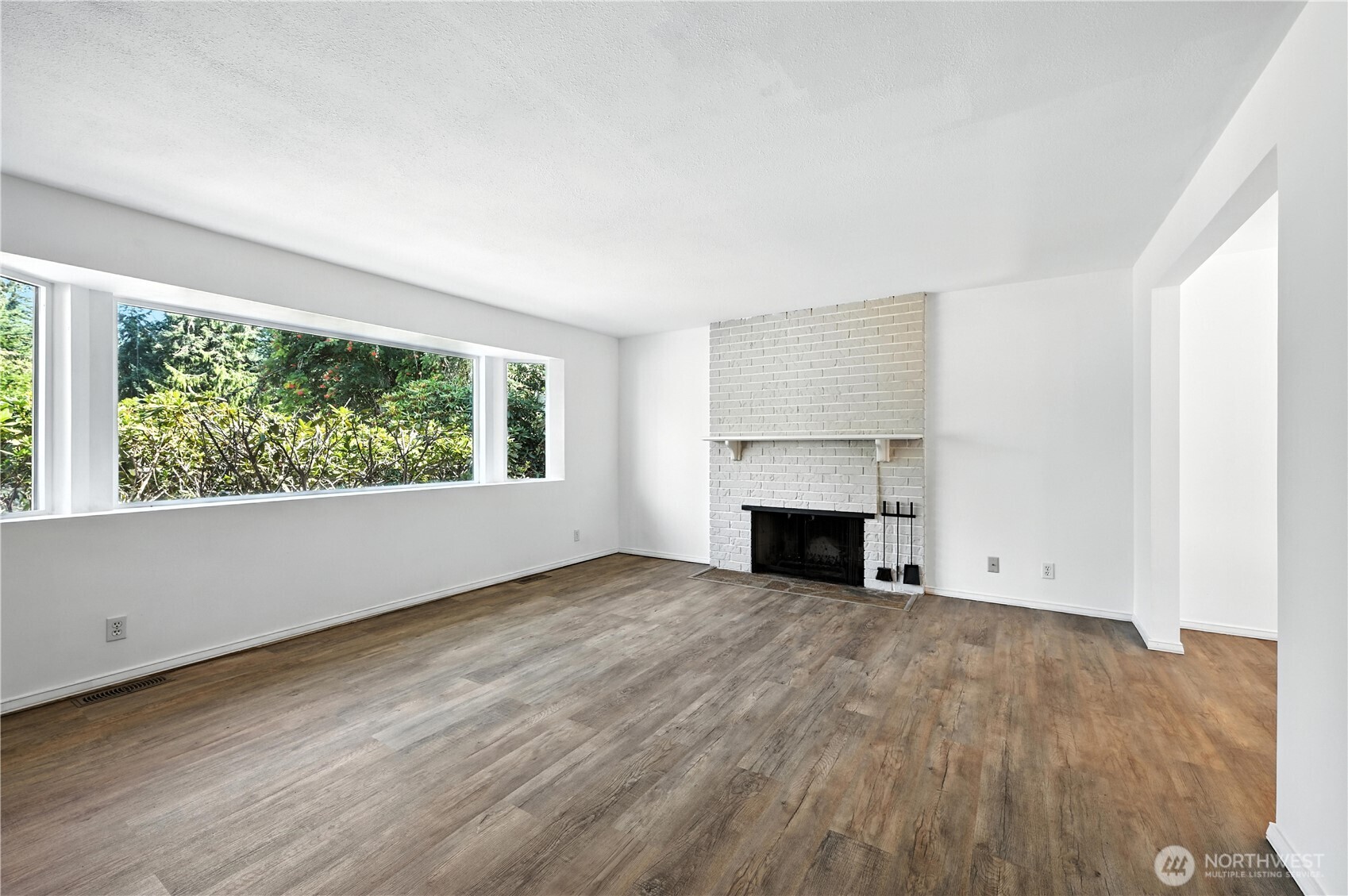 Vacant living room with a white brick fireplace, wood-look floors, and wide windows facing greenery.