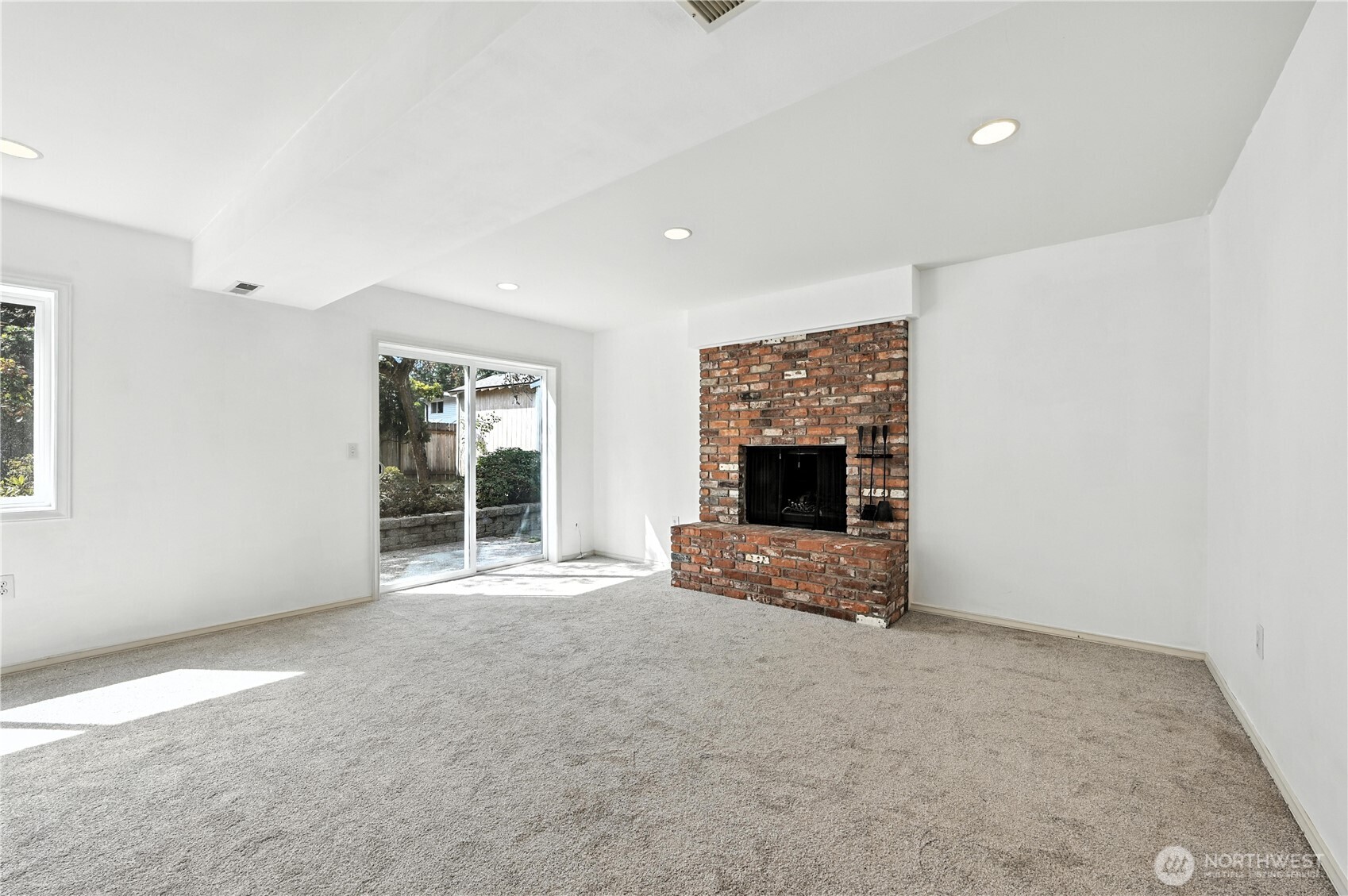 Empty living room with a brick fireplace, light carpet, and sliding glass door to the yard.