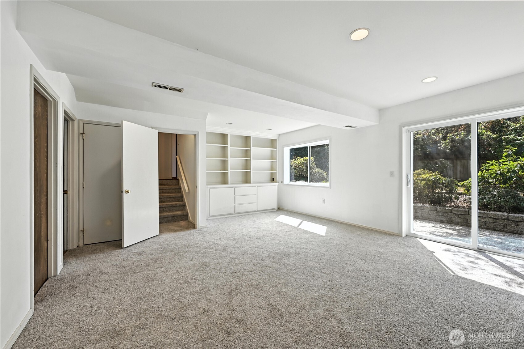 Empty carpeted room with built-in shelves, stairs, and sliding glass doors to a patio.