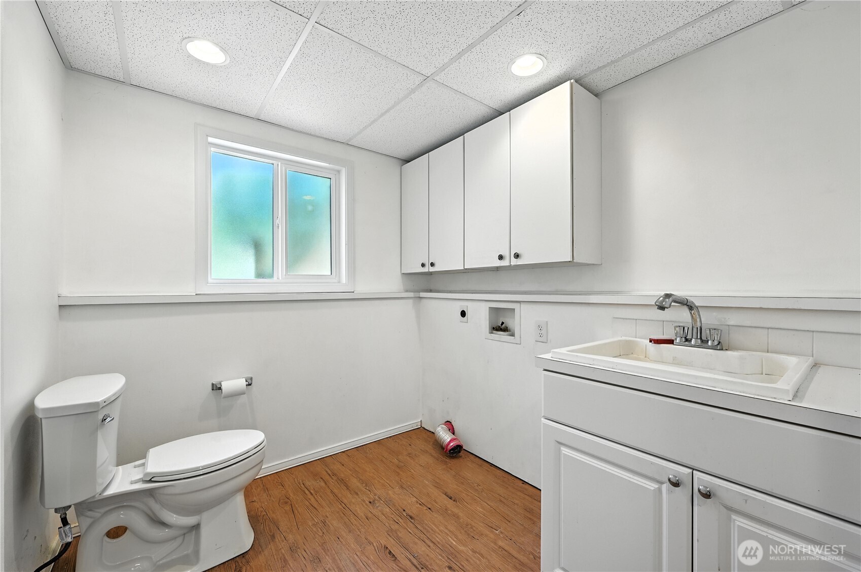 Utility bathroom with a toilet, deep sink, white cabinets, and wood-look flooring.