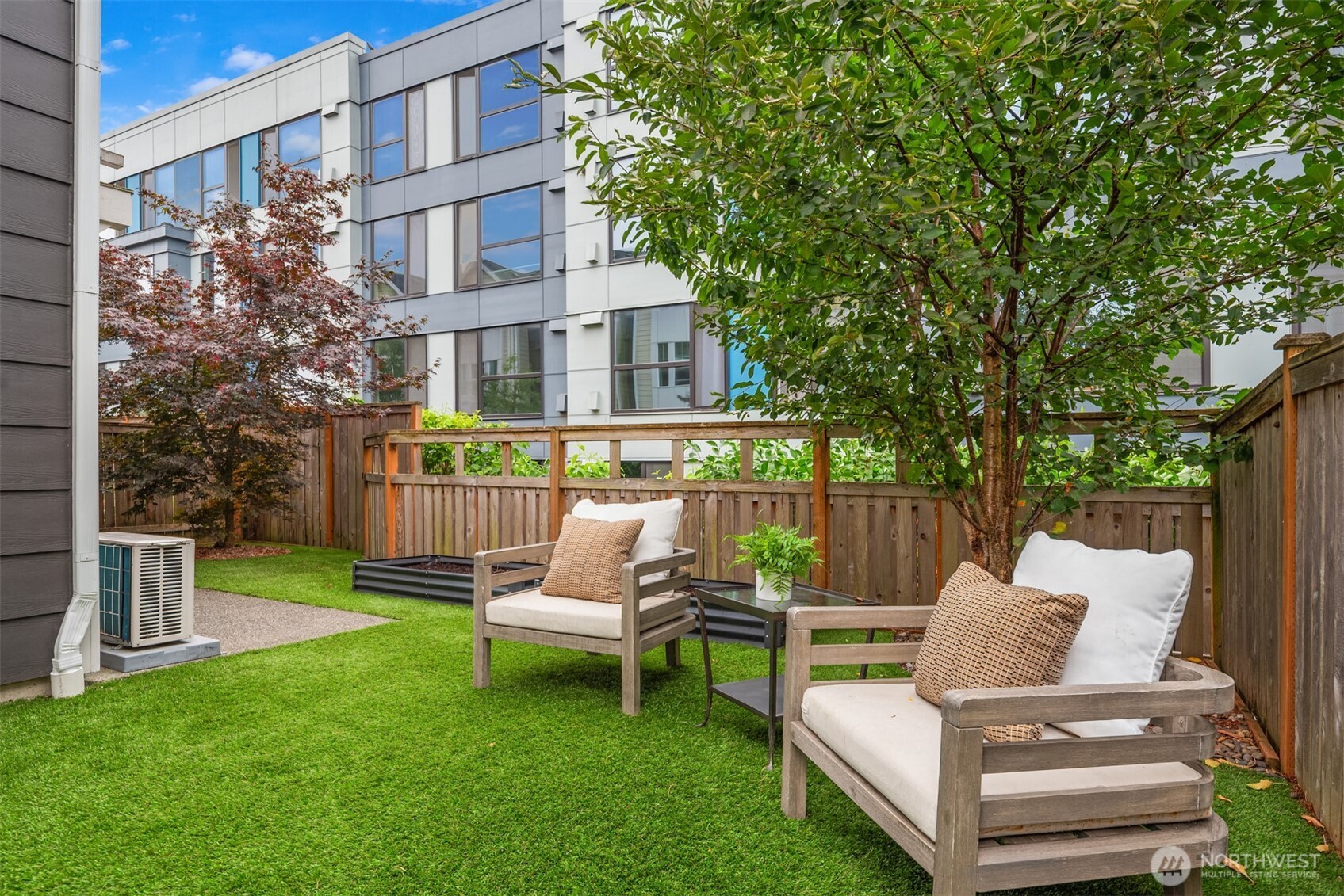 Private patio with cushioned wooden chairs, a green lawn, mature trees, and a modern building beyond the fence.