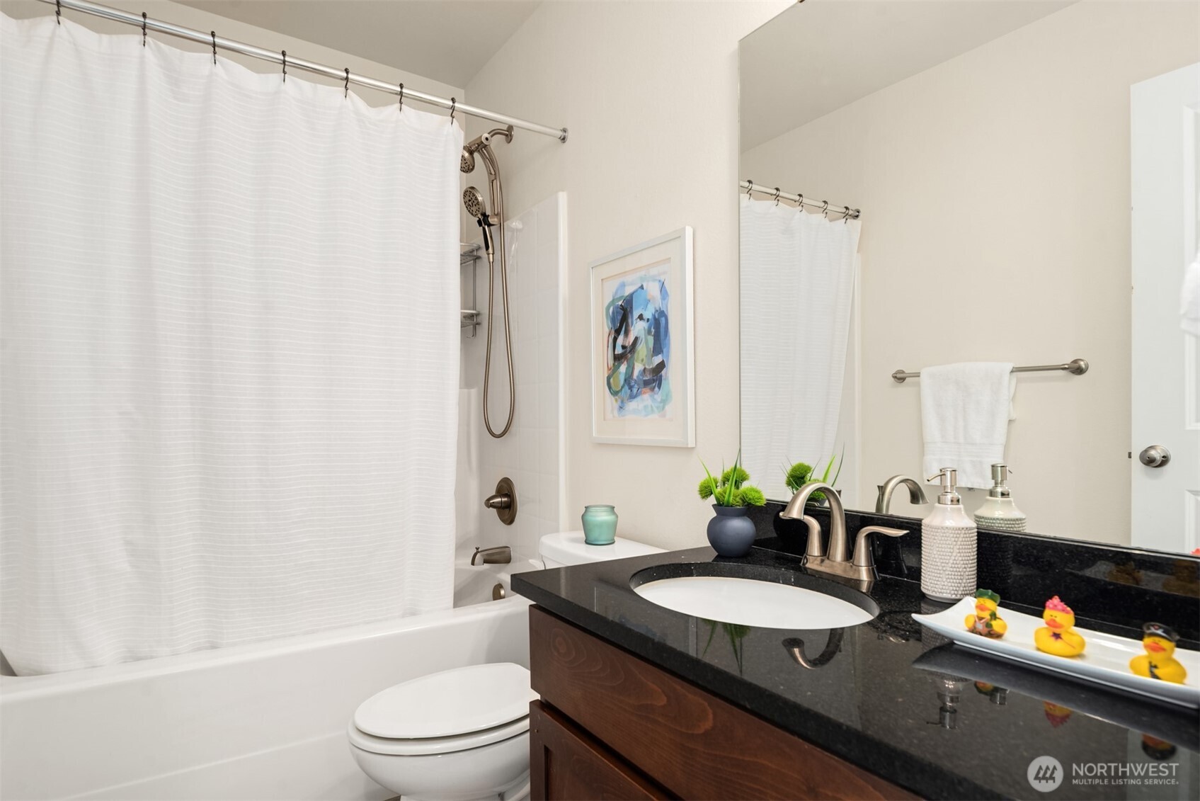 Bathroom with a white tub and shower, framed abstract art, and a dark granite vanity decorated with rubber ducks.