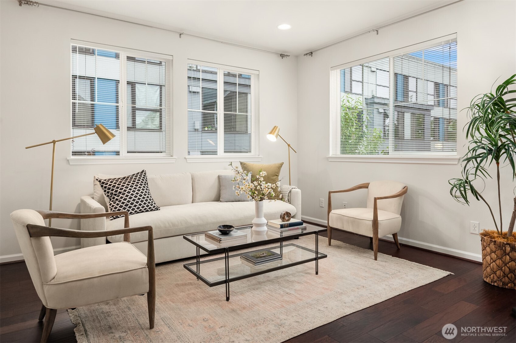 Staged living room with a white sofa, two chairs, and large windows overlooking neighboring buildings.