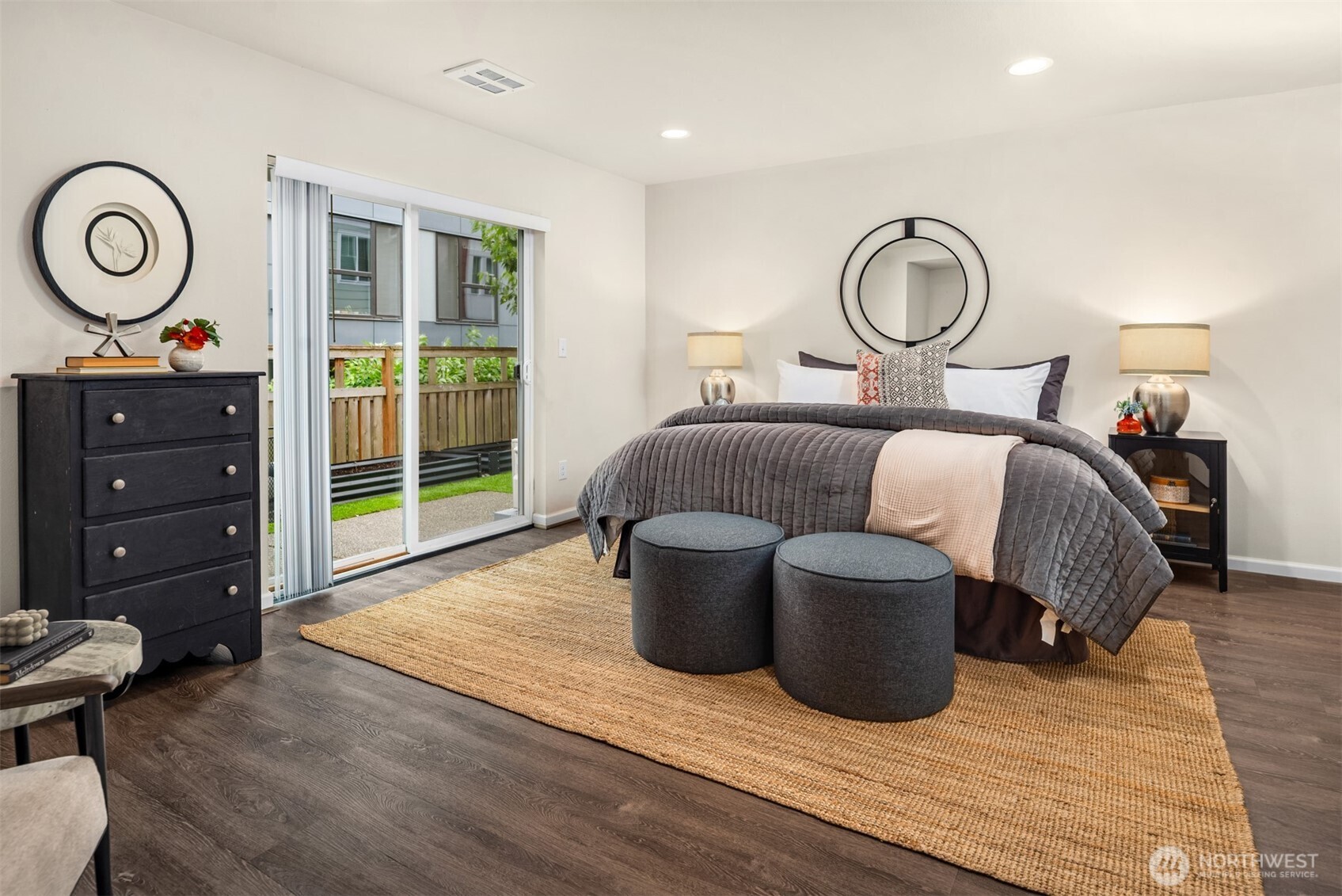 Staged bedroom with a gray-quilted bed, round ottomans, and sliding glass door to a patio.