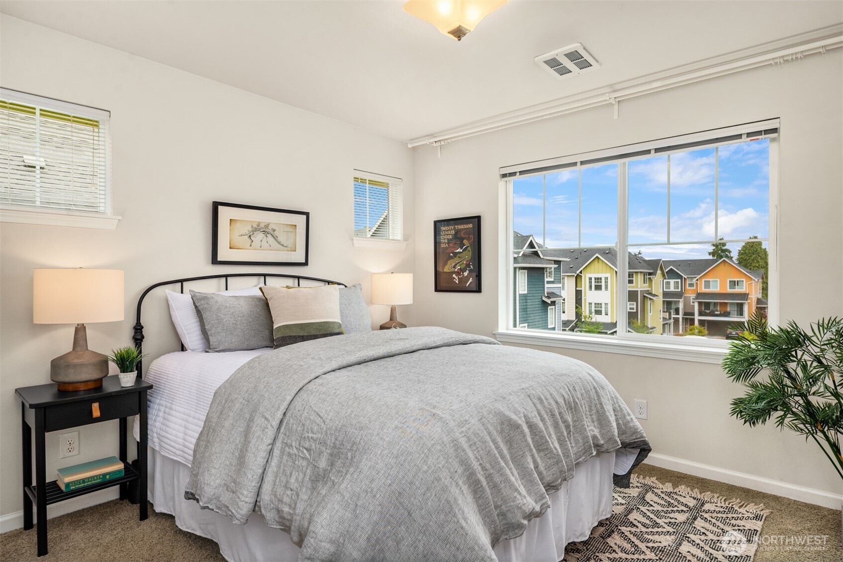 Staged bedroom with gray bedding, framed art, and a large window overlooking colorful townhomes.