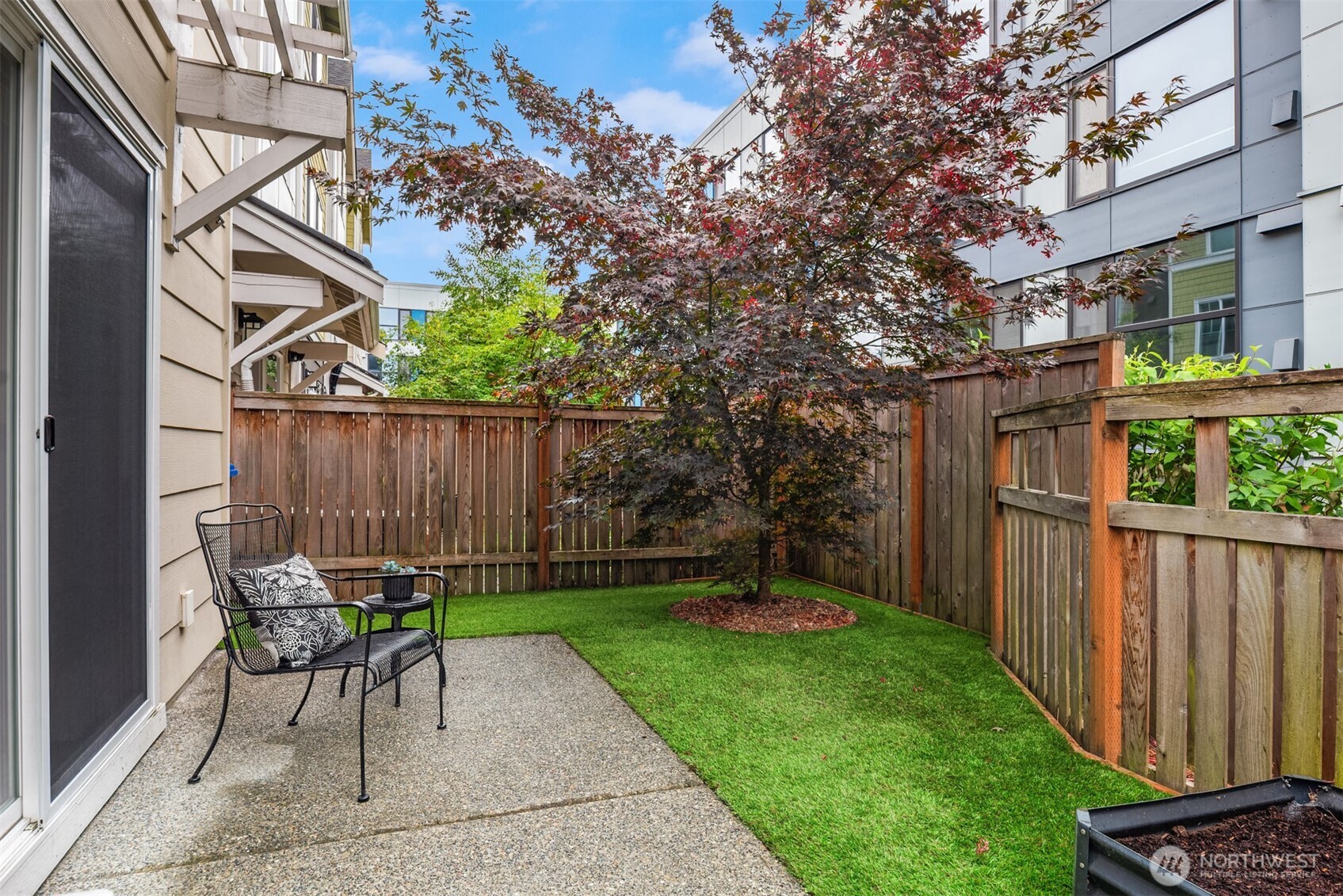 Private condo patio with a fenced lawn, metal chairs, and a red-leaved Japanese maple.