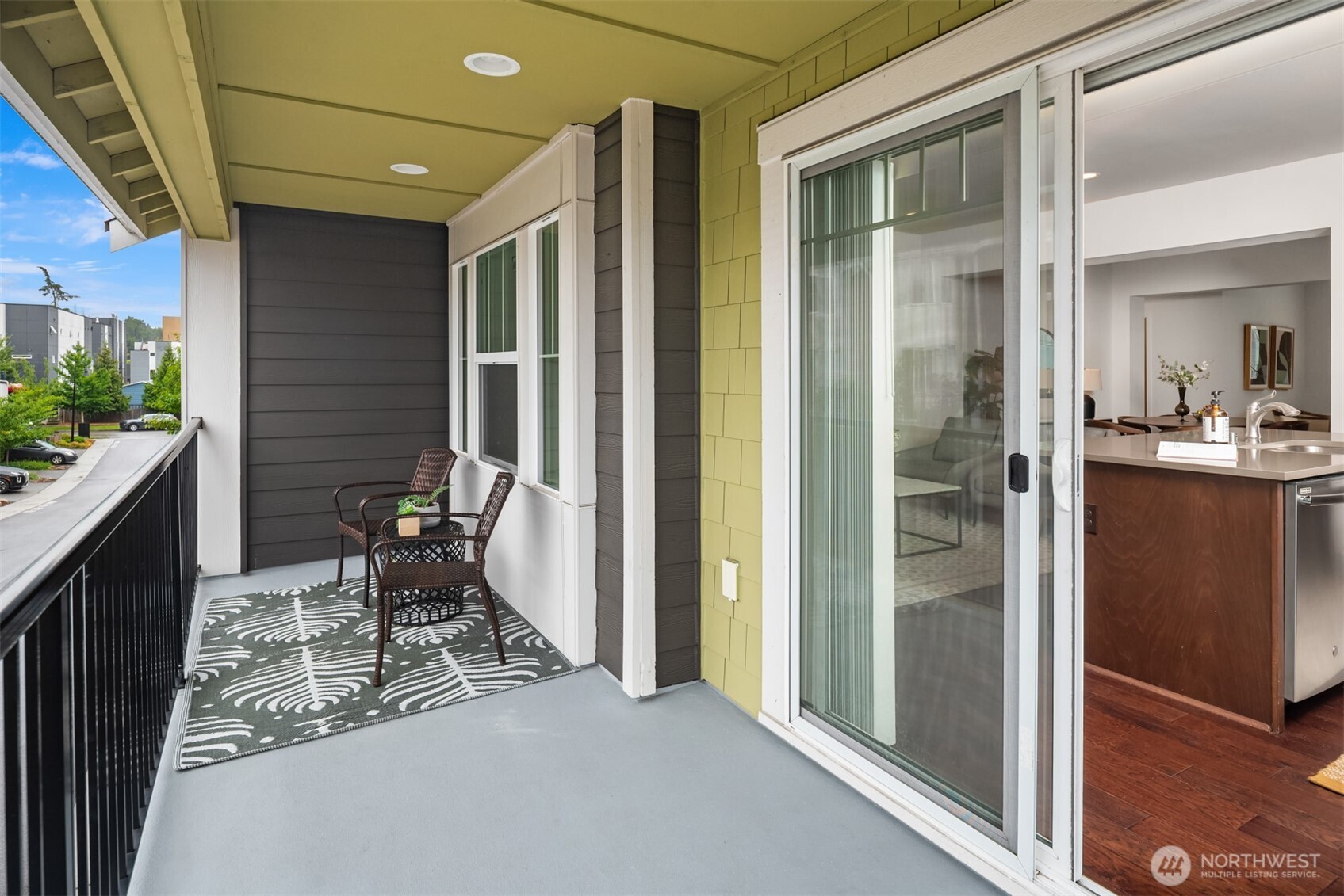 Covered apartment balcony with wicker chairs and a patterned rug beside sliding doors to the kitchen.