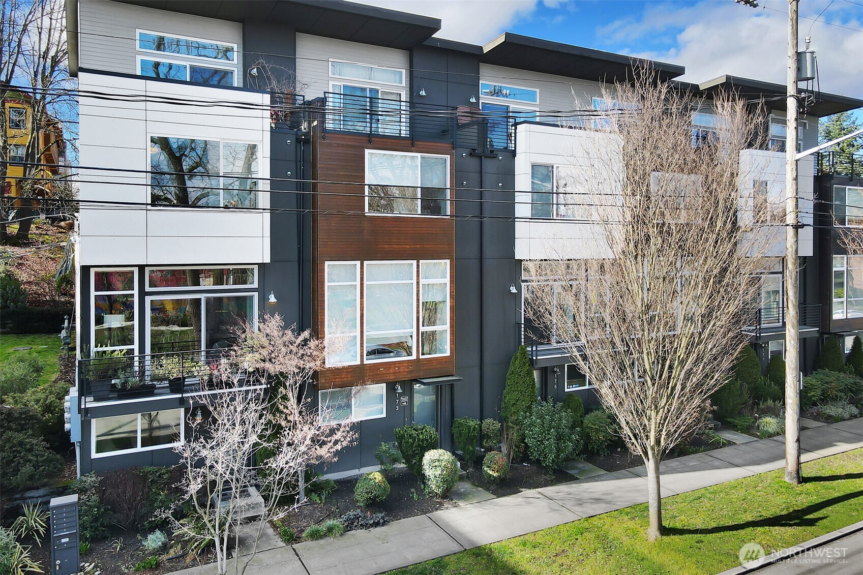 Modern multi-unit building with dark gray siding, wood accents, balconies, and leafless street trees.