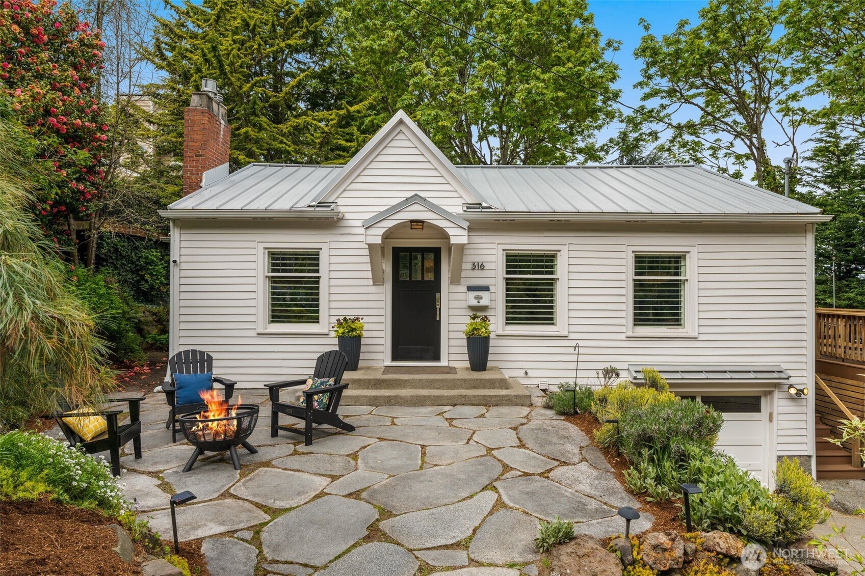 White house with a metal roof, stone patio, Adirondack chairs, and a fire pit.