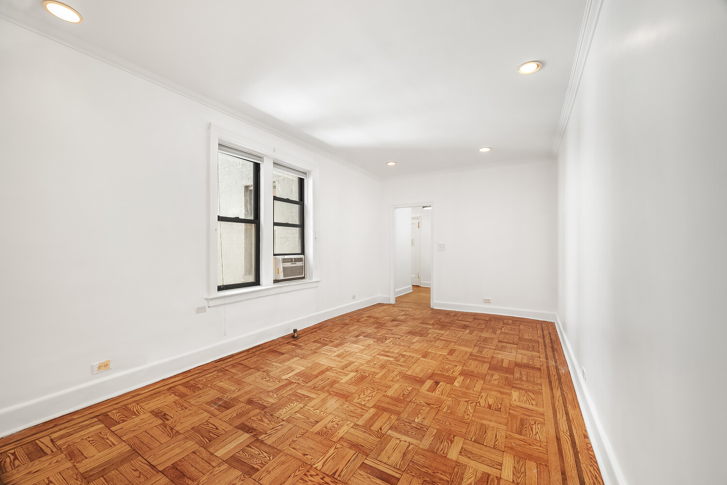 Empty room with parquet floors, white walls, and two windows, one fitted with an air conditioner.