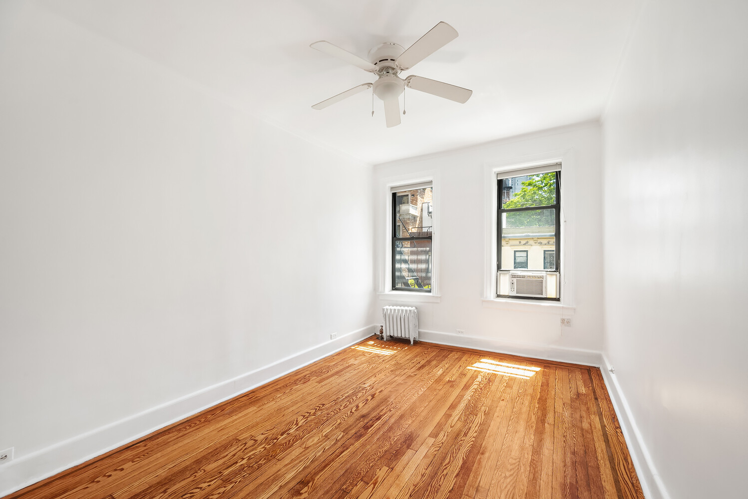 Empty bedroom with polished wood floors, two windows, a radiator, and a ceiling fan.