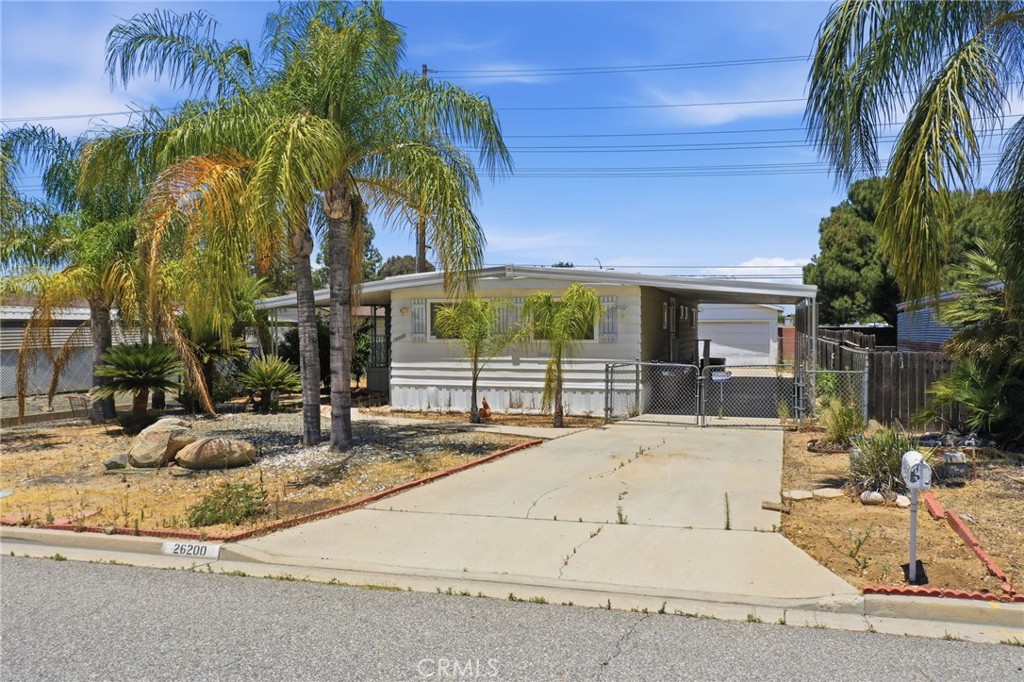 Single-story manufactured home with a gated driveway, palm trees, and the number 26200 on the curb.