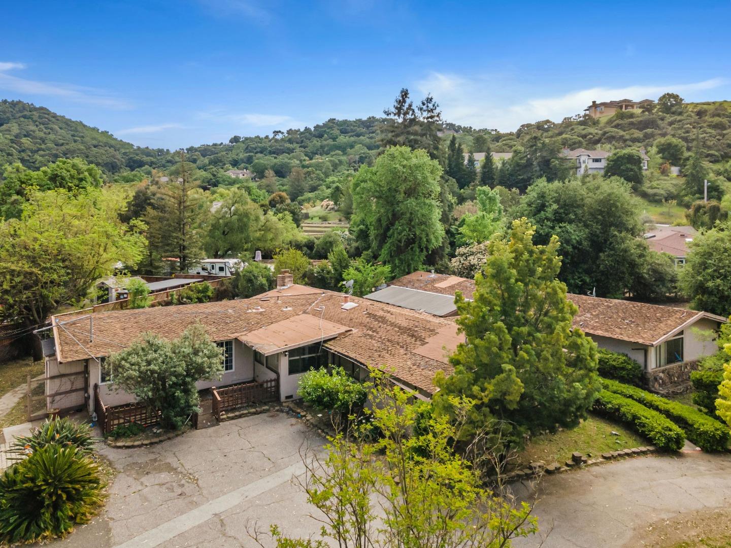 Aerial view of a tan-roofed home among trees and neighboring houses in a green hillside neighborhood.