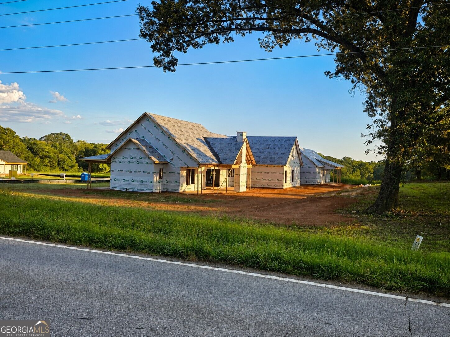 House under construction beside a road, with wrapped walls, exposed framing, and a large tree.