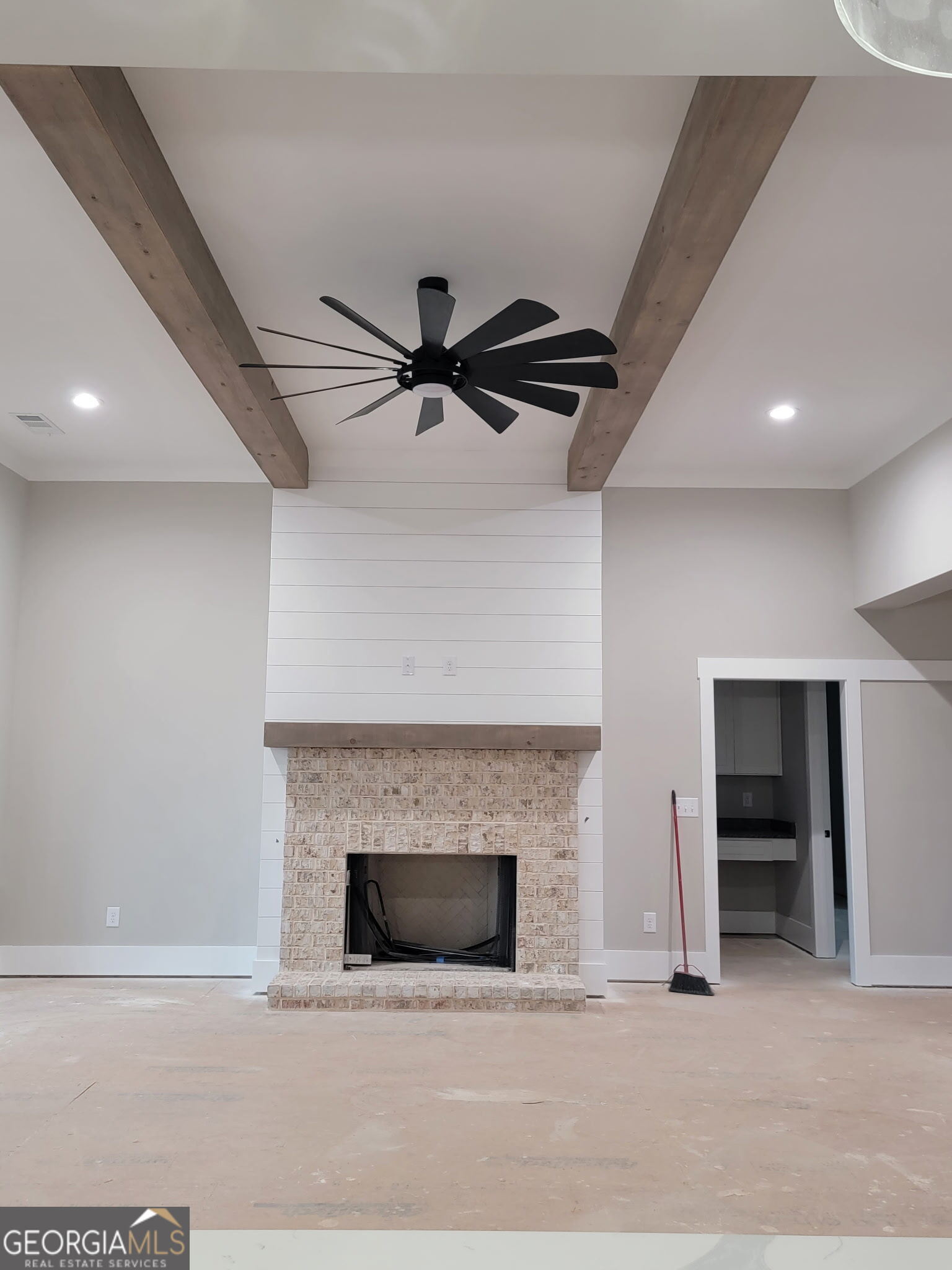 Living room with a brick fireplace, shiplap surround, exposed ceiling beams, and black ceiling fan.