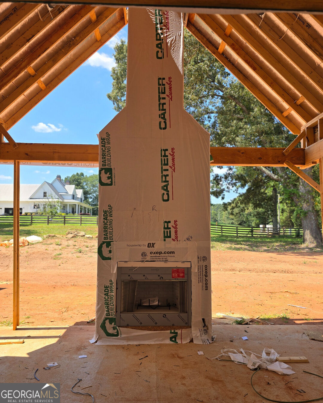 Fireplace and exposed timber framing in an unfinished structure, with a white house and fenced yard beyond.