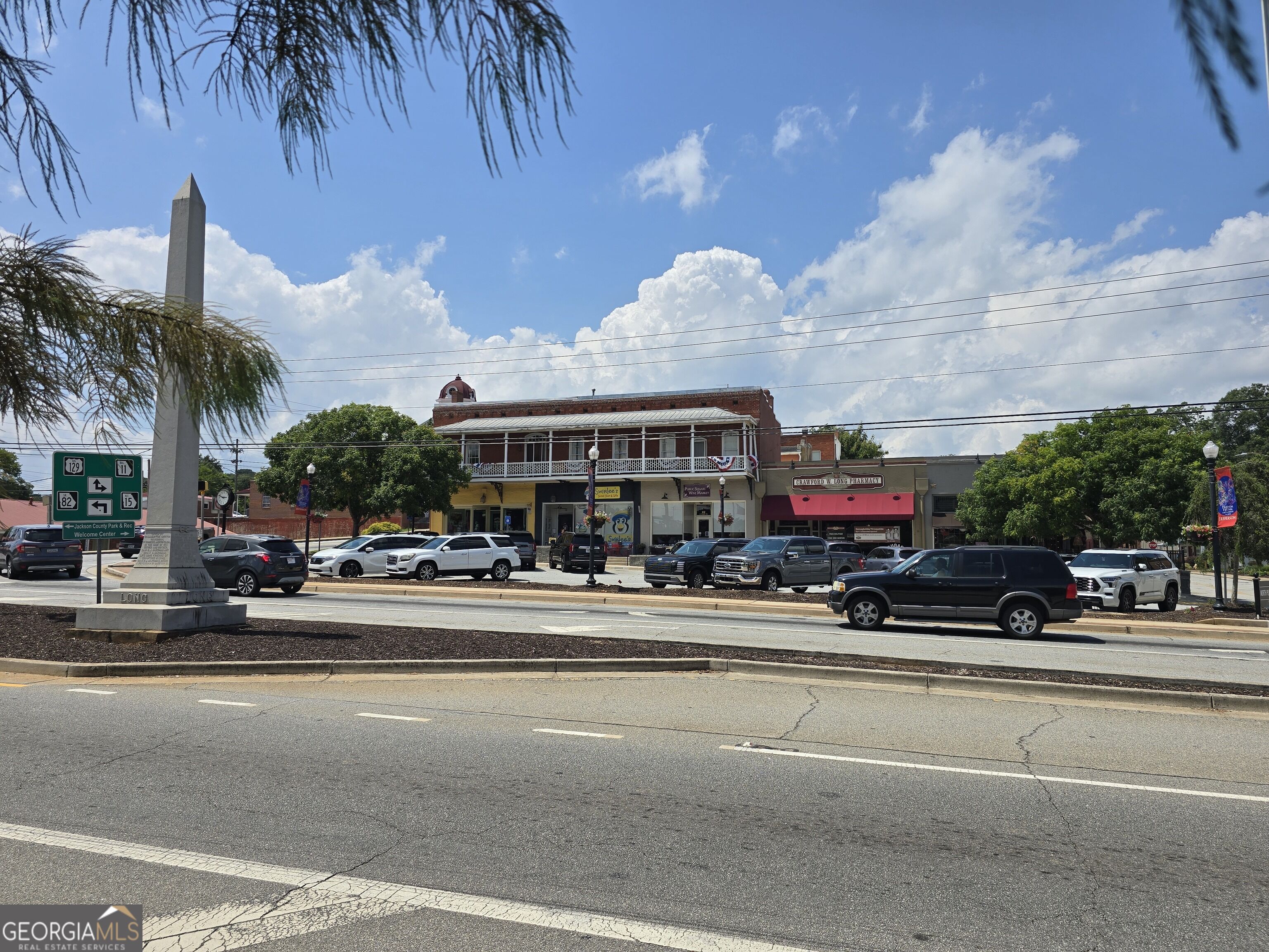 Downtown storefronts and parked cars across the street from a tall stone obelisk.