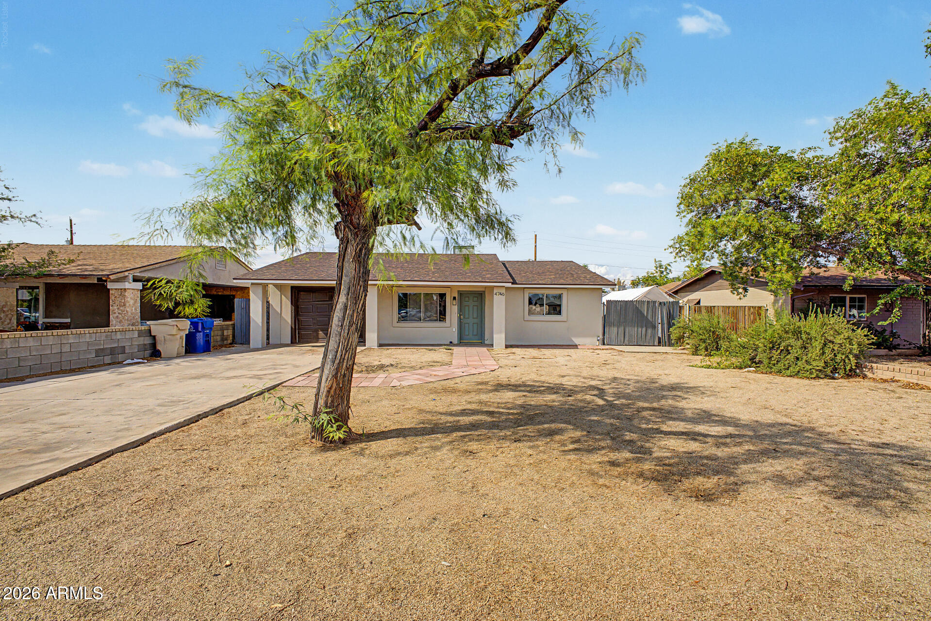 Single-story stucco home with a blue-green door, dry gravel yard, large shade tree, and concrete driveway.