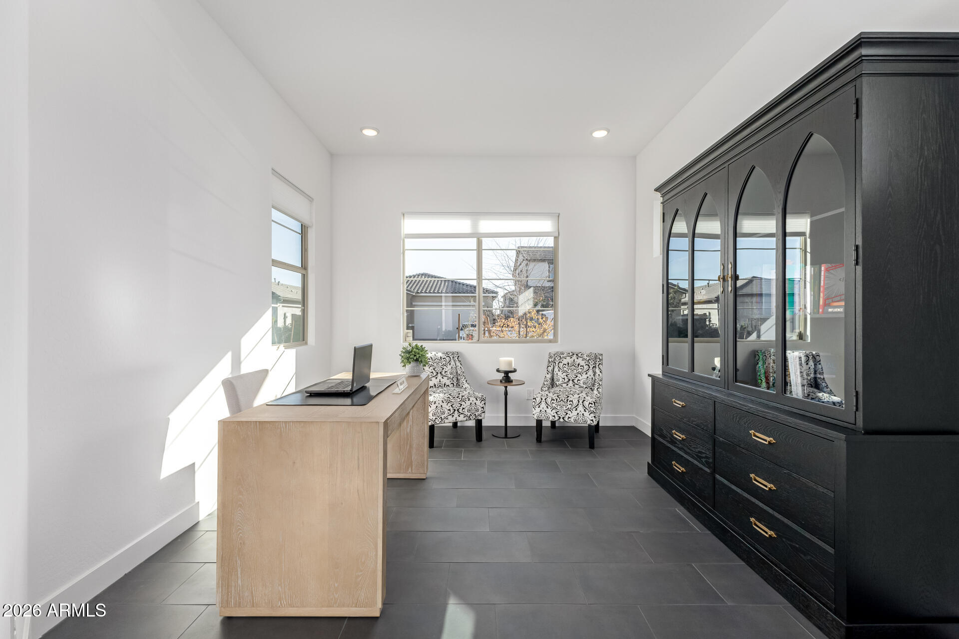Bright home office with a light wood desk, patterned armchairs, and a tall black glass-front cabinet.