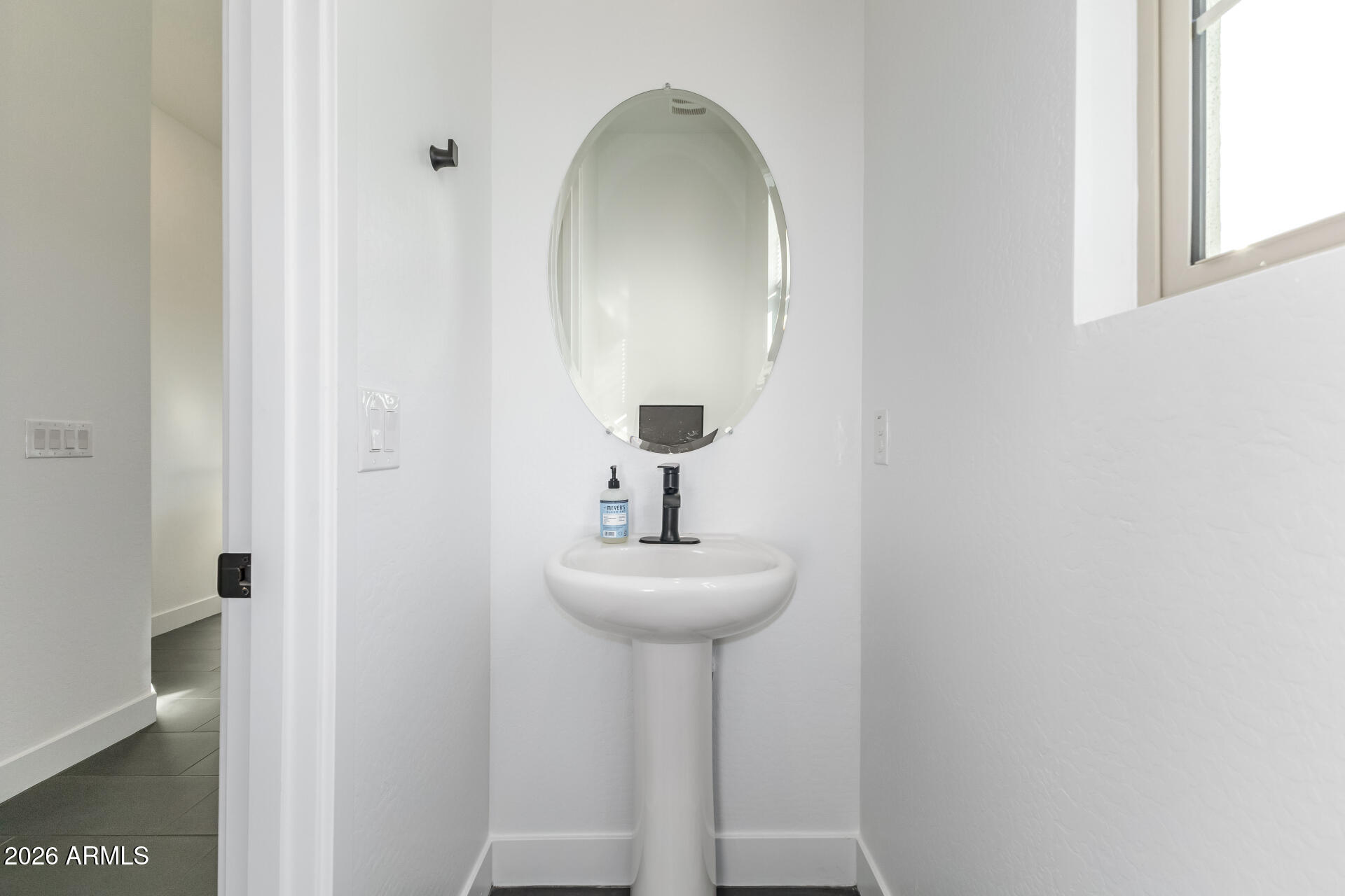 Bathroom pedestal sink with black faucet, oval mirror, and window.