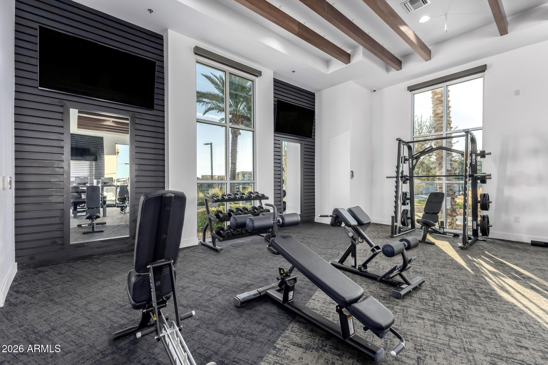 Fitness room with weight machines, dumbbells, tall windows, and exposed ceiling beams.