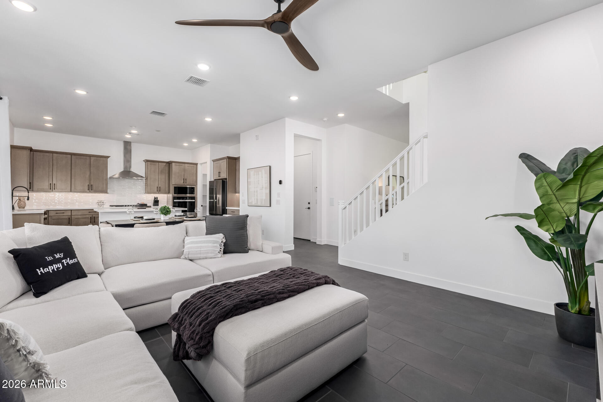 Open-plan living room with a white sectional, dark tile floors, kitchen island, and staircase.
