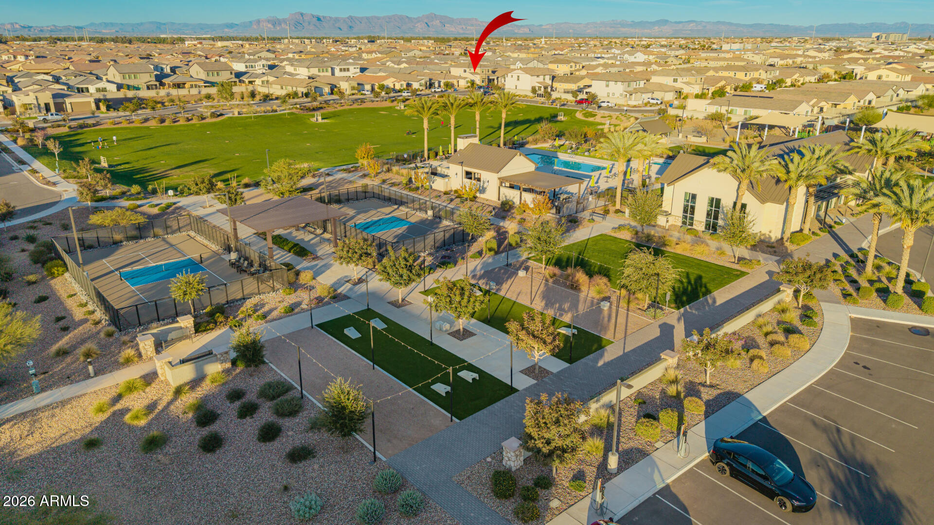 Aerial view of the home marked by a red arrow amid community parks, courts, a pool, and distant mountains.