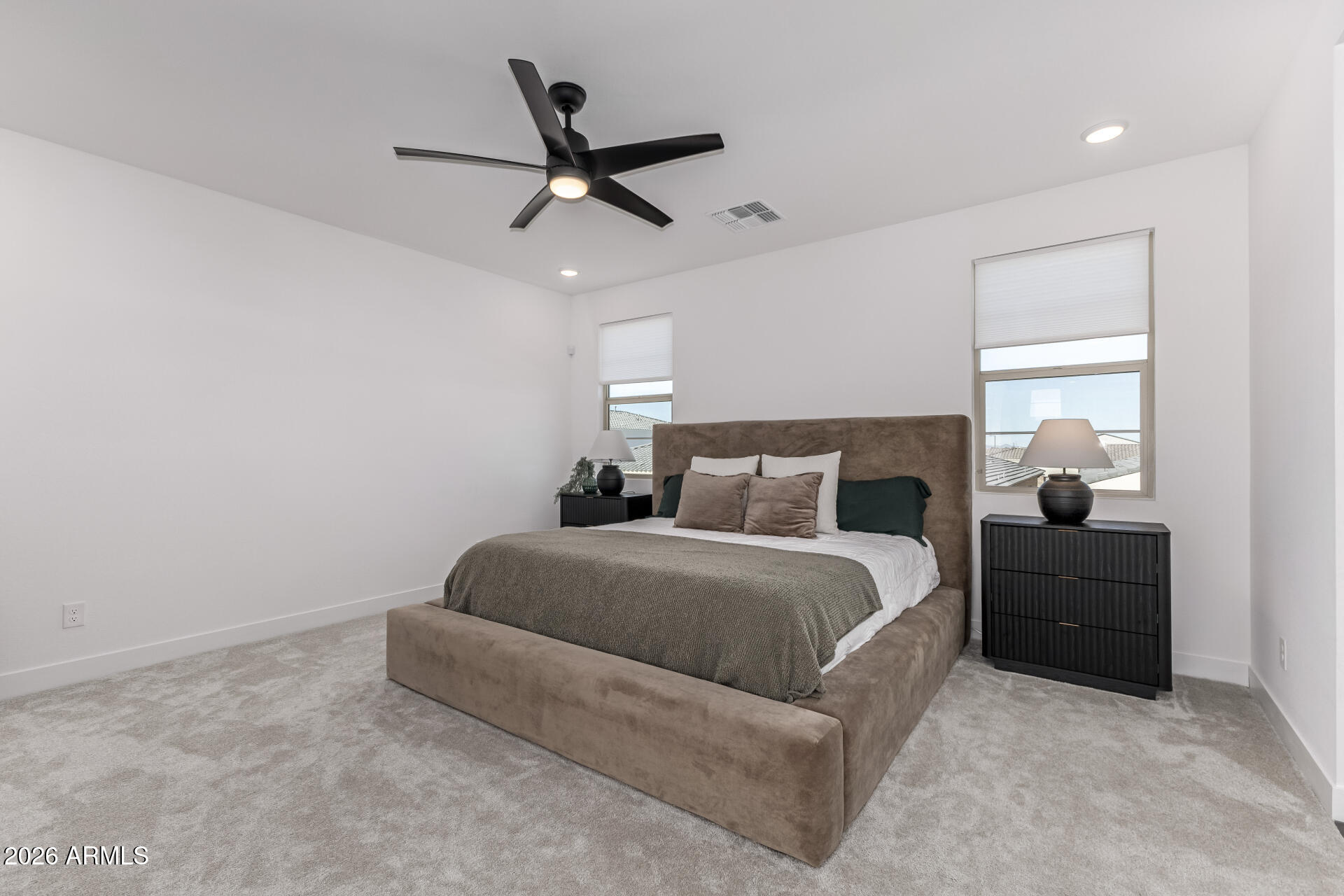 Staged bedroom with an upholstered bed, gray carpet, black nightstands, and a ceiling fan.