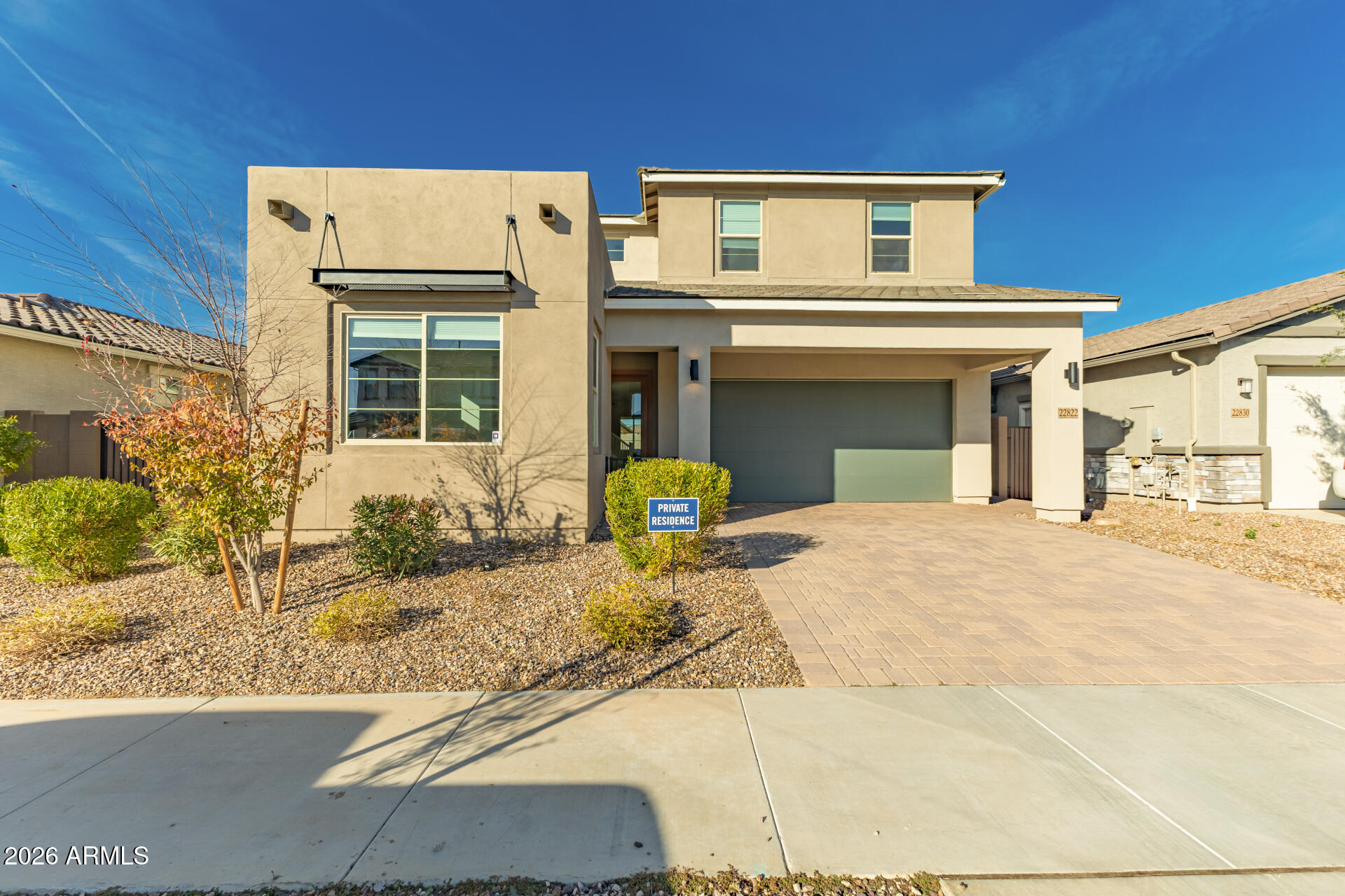 Two-story beige stucco home with a double garage, paver driveway, and gravel landscaping.