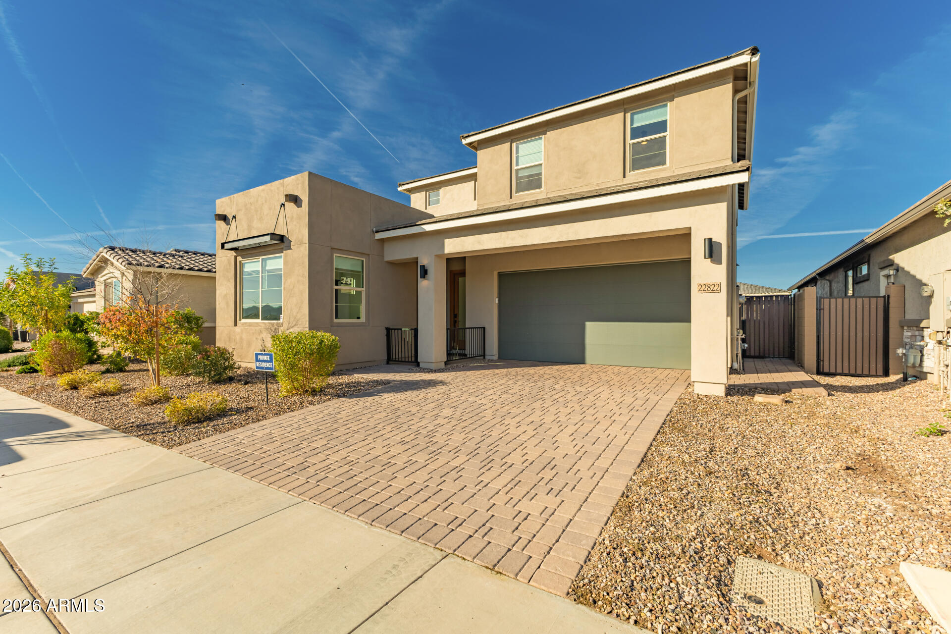 Two-story beige stucco home with a gray garage, brick-paver driveway, and desert landscaping.