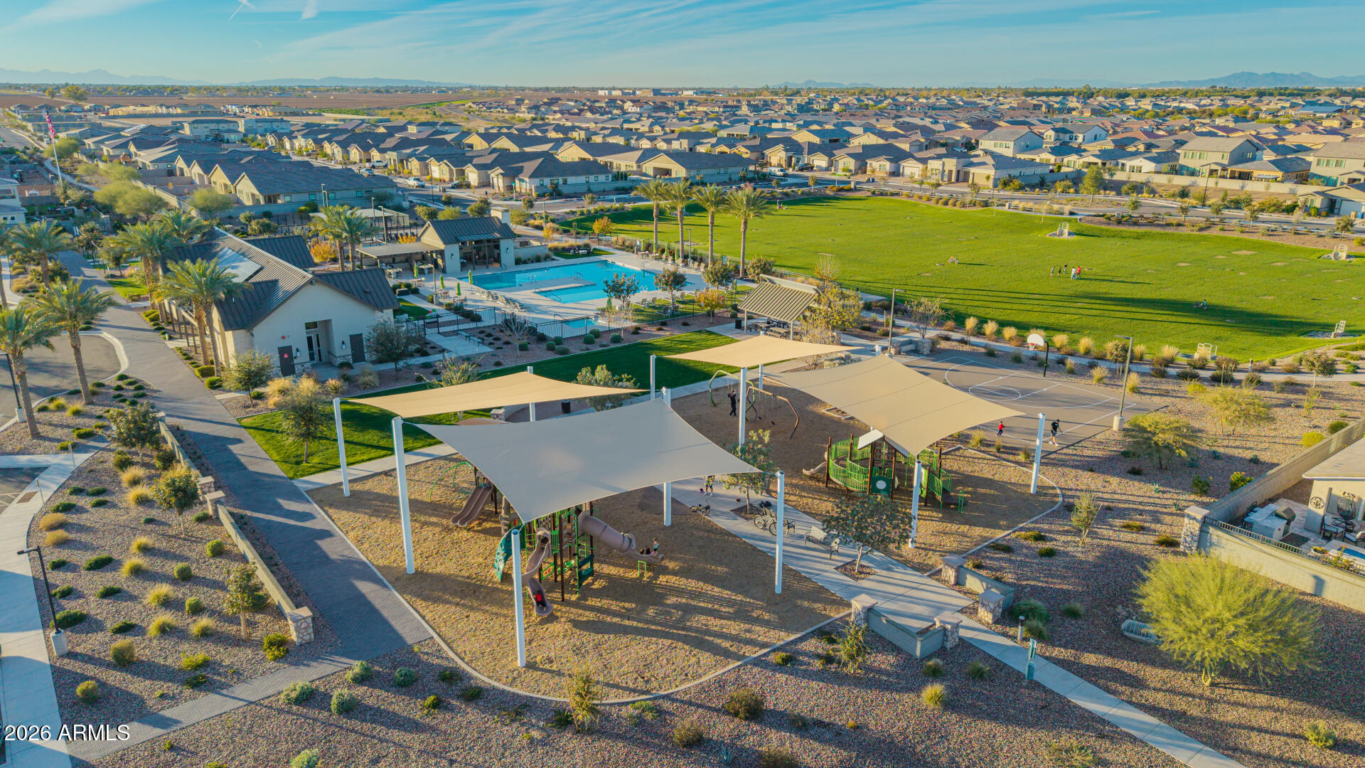 Aerial view of a neighborhood park with a pool, shaded playground, sports courts, and a large grassy field.