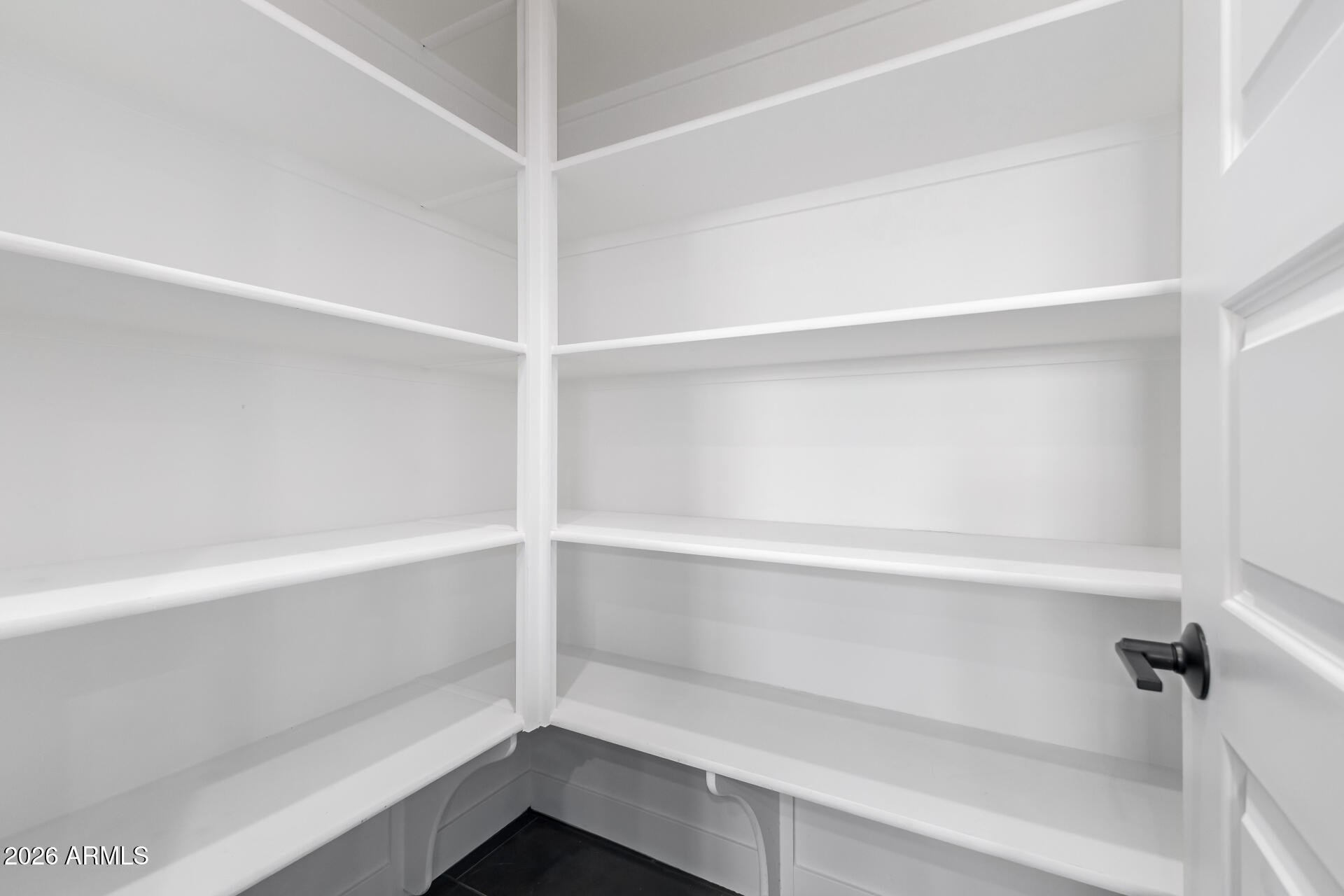 Empty walk-in pantry with white wraparound shelves and a dark tile floor.