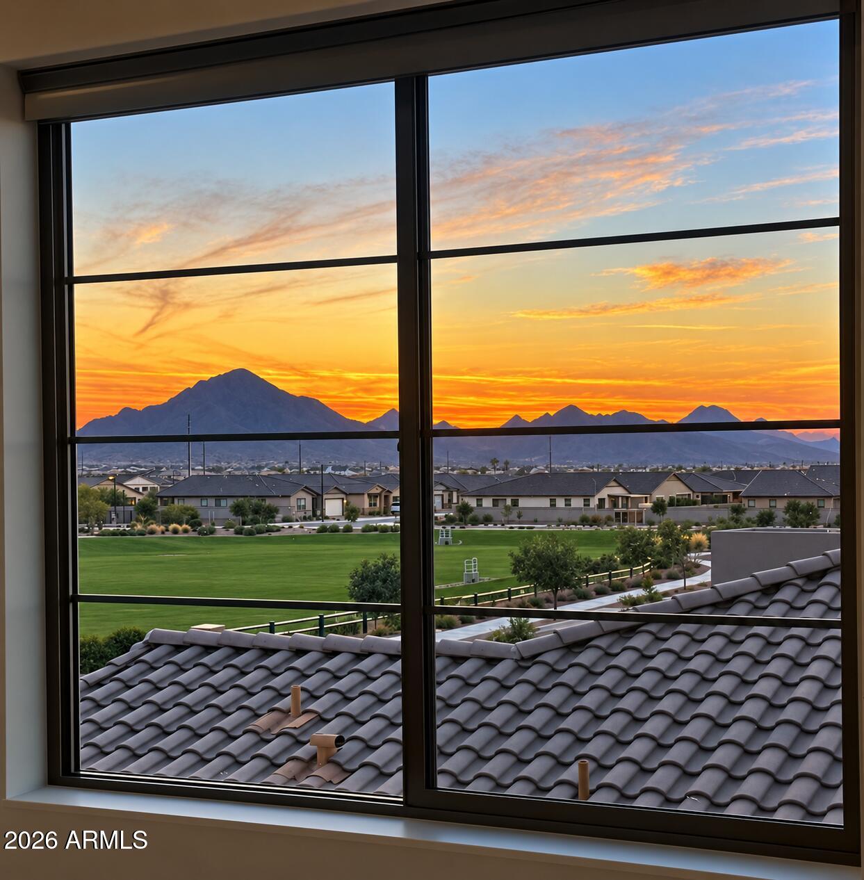 Mountain sunset and neighborhood view through a large window, with a green lawn and tiled rooftops below.