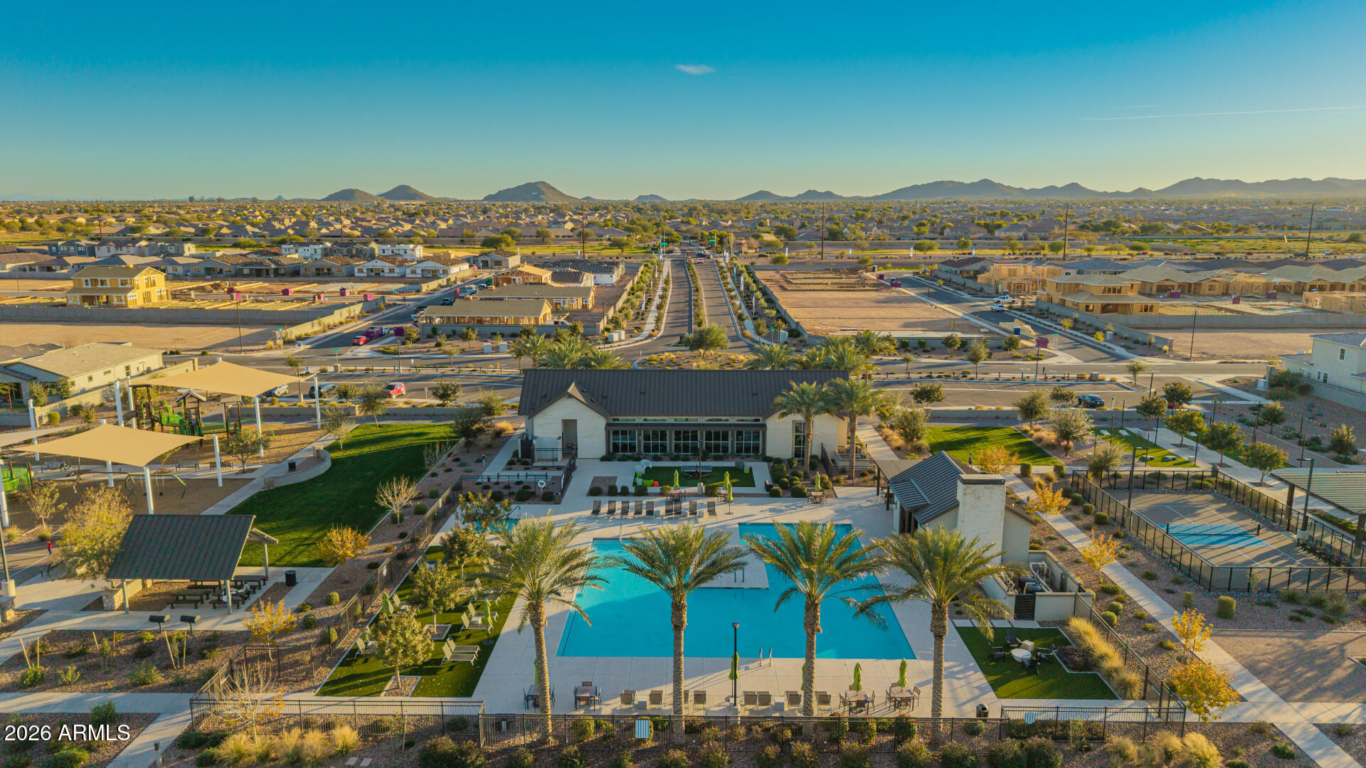 Aerial view of a community pool, clubhouse, sports courts, and new homes with mountains beyond.