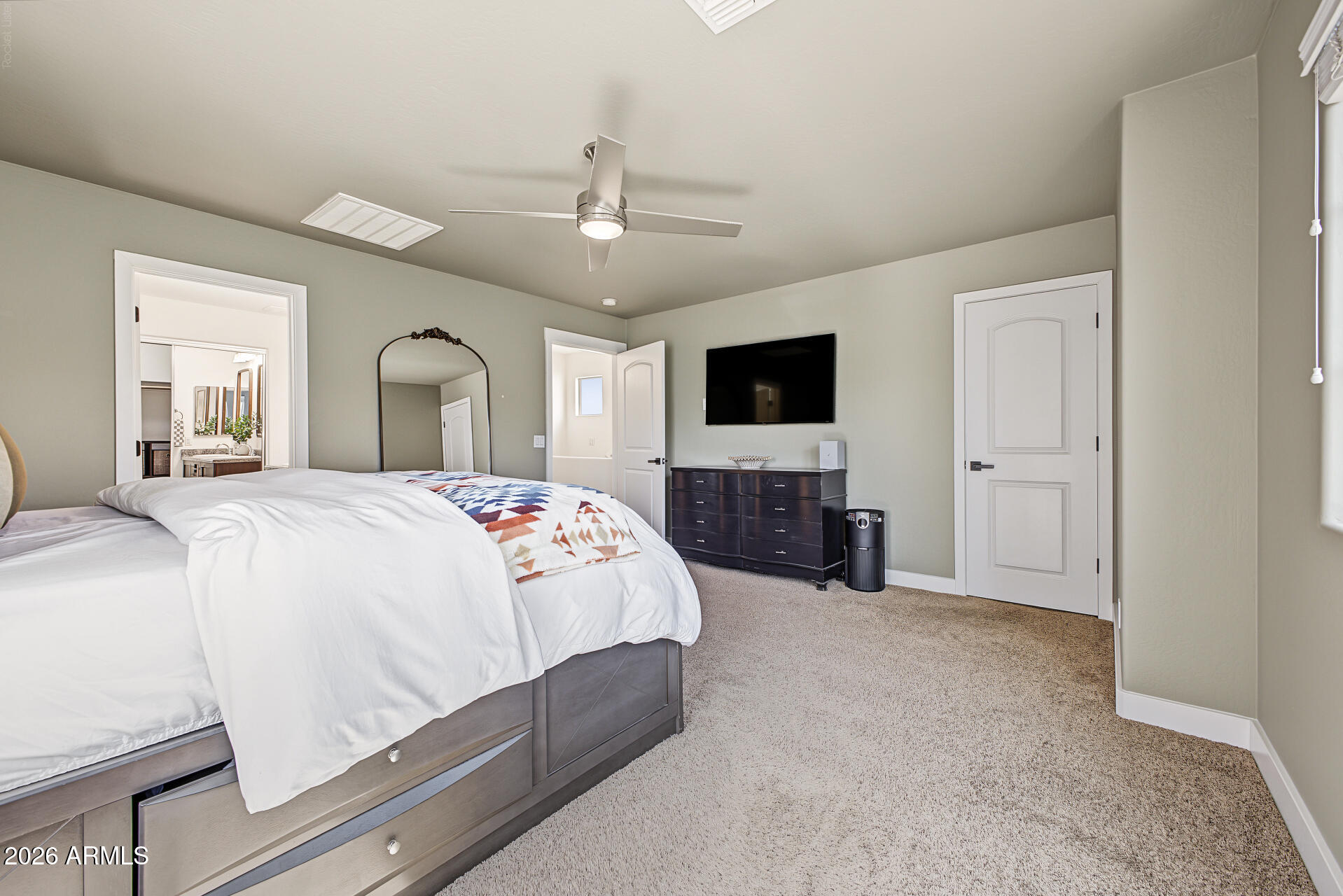 Furnished bedroom with a large white bed, dark dresser, wall-mounted television, and ceiling fan.