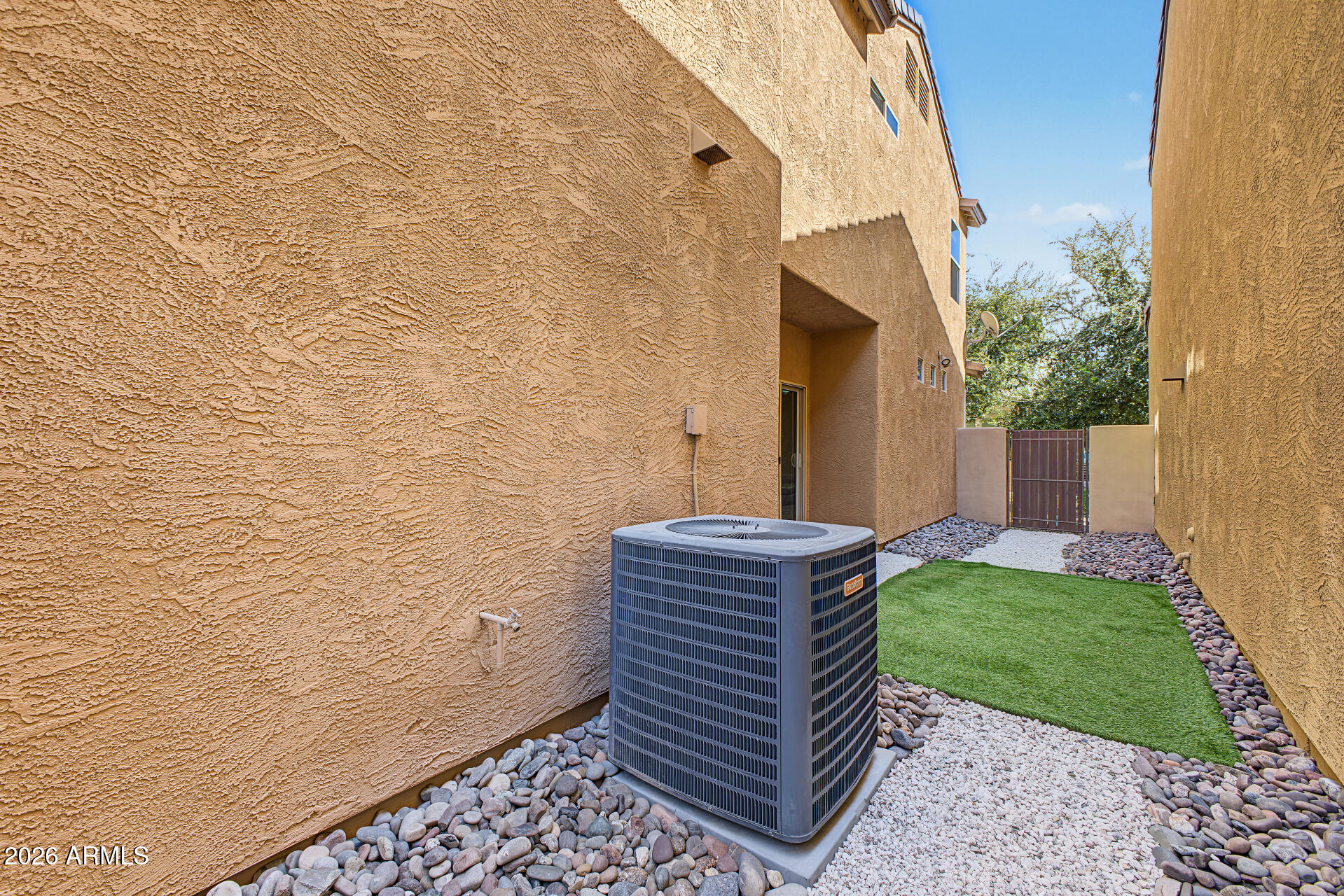 Side yard with an air-conditioning unit, artificial grass, decorative rocks, and a metal gate.