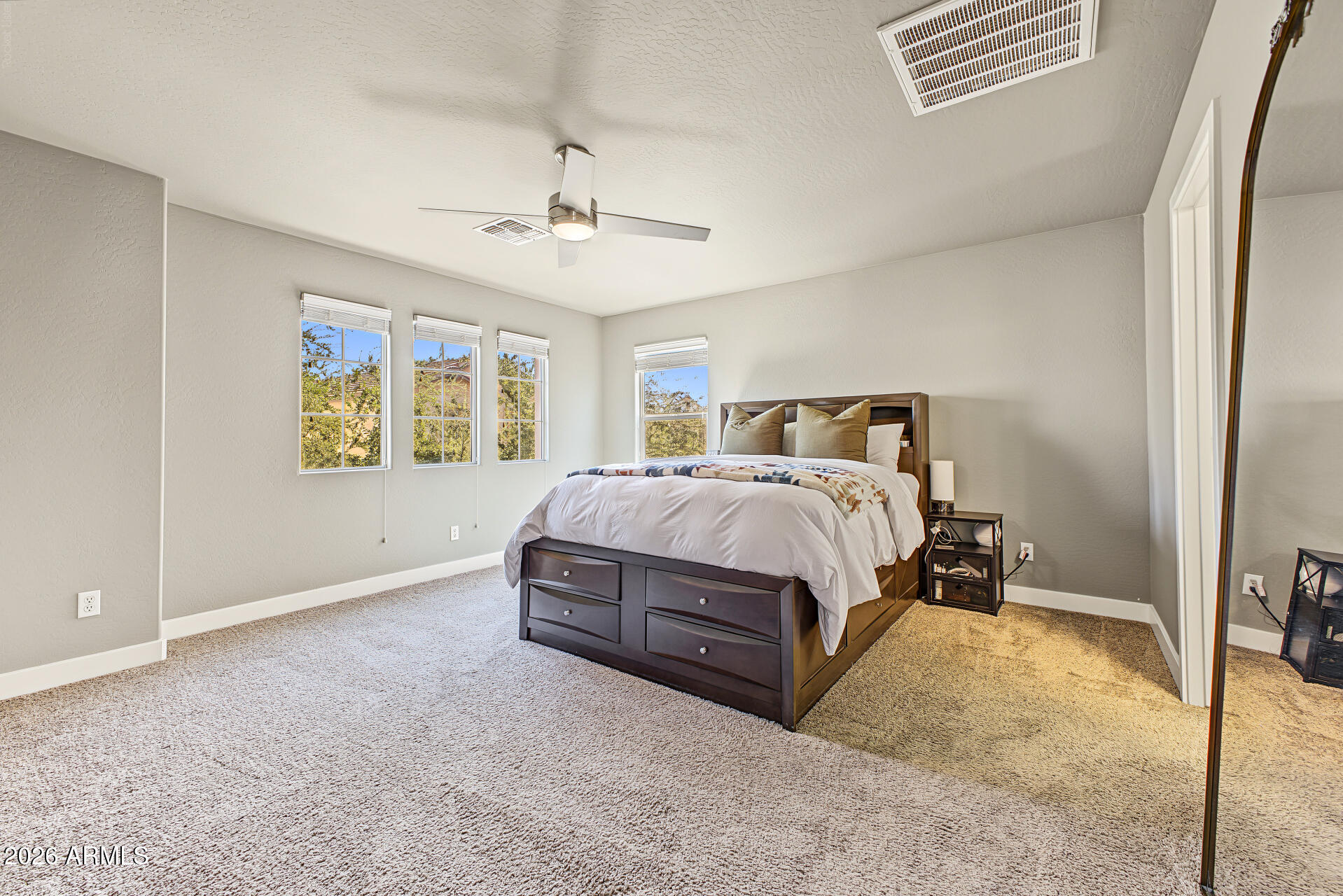 Furnished bedroom with a dark wood bed, ceiling fan, and multiple windows overlooking trees.