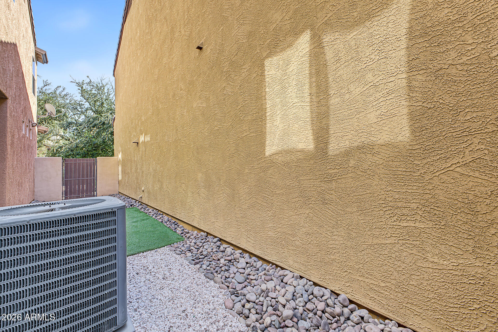 Side yard with a large air-conditioning unit, gravel landscaping, artificial turf, and a metal gate.