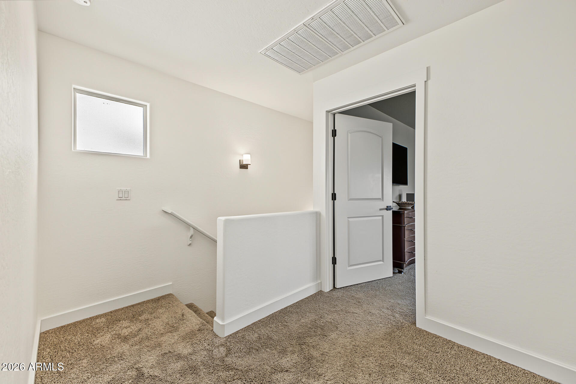 Carpeted upstairs landing with a stairwell, white half-wall, and open door to a room with a dresser and television.