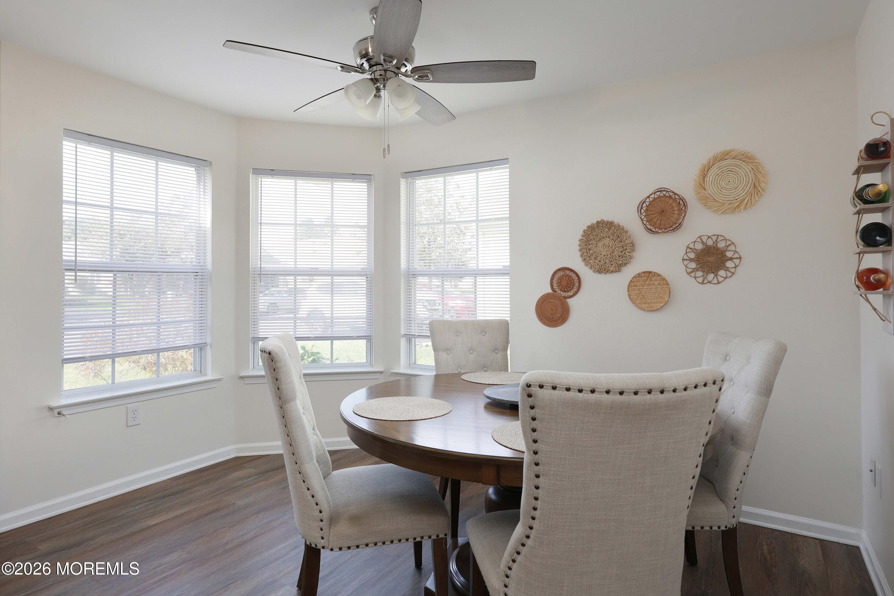 Dining room with a round wood table, upholstered chairs, three bright windows, and woven wall decor.